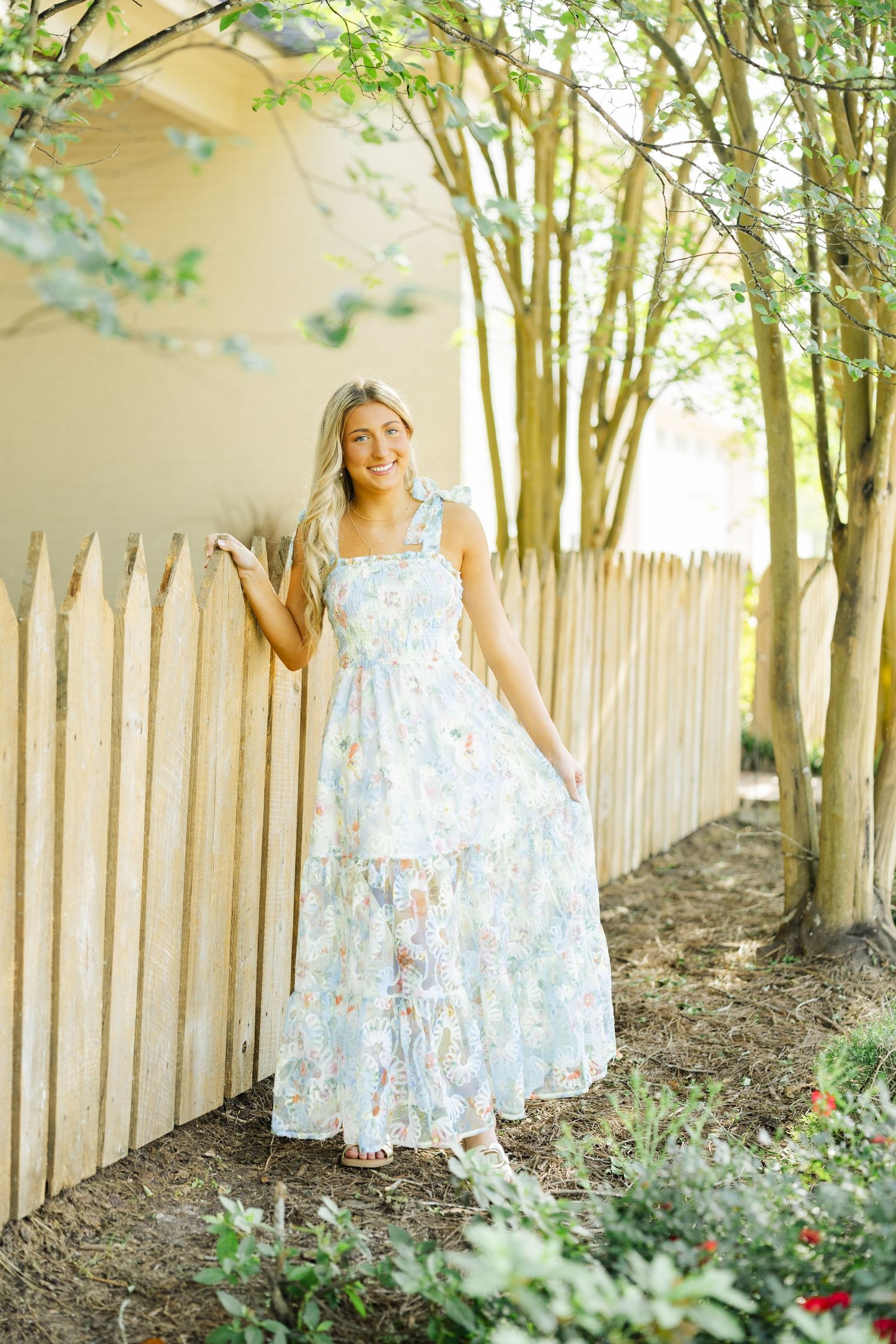 Blonde high schooler in floral maxi dress leans against a wooden picket fence in a garden setting.