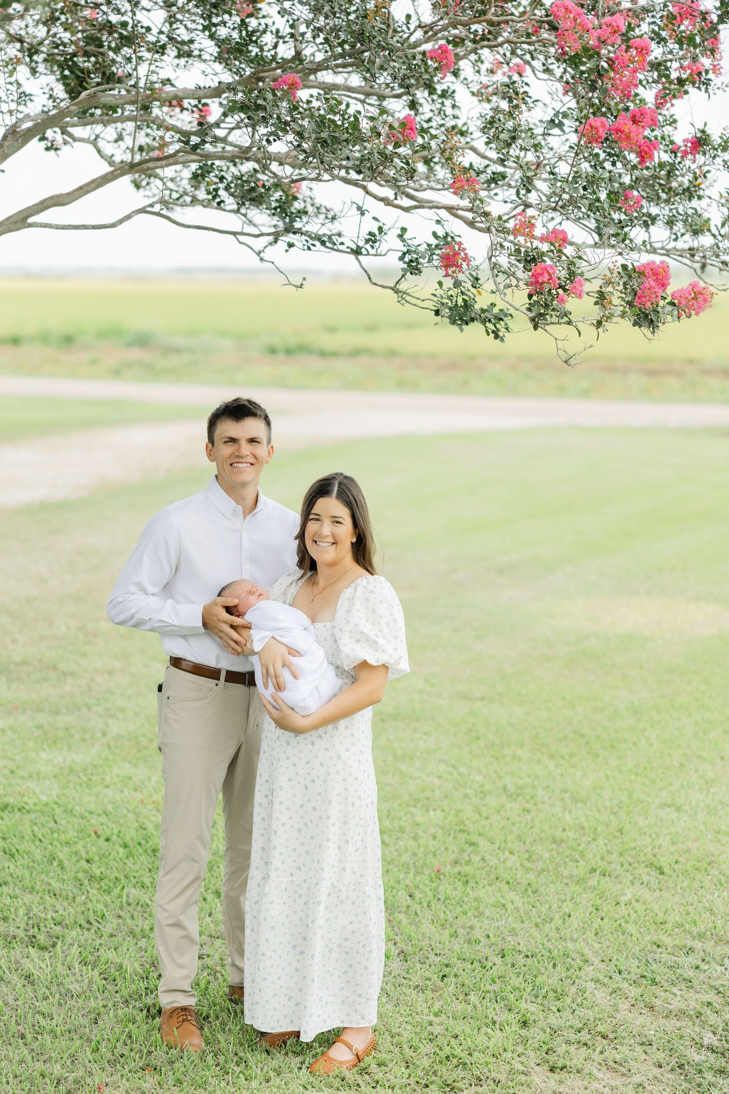 Smiling new parents stand under blooming pink flower tree holding their swaddled newborn baby outdoors.