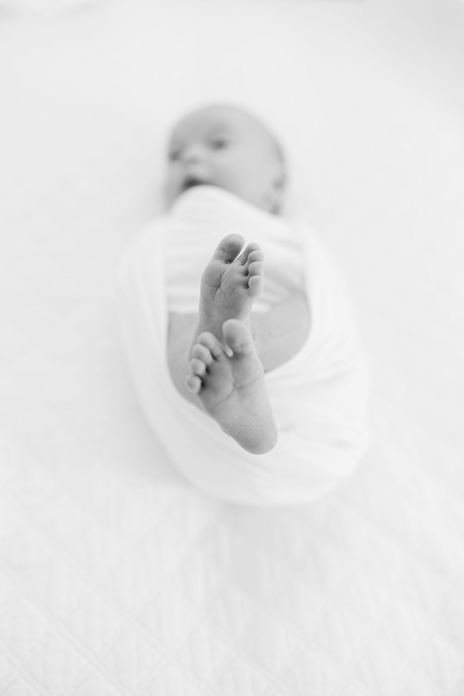 Black and white close-up of newborn baby feet peeking from white swaddle wrap on soft blanket.