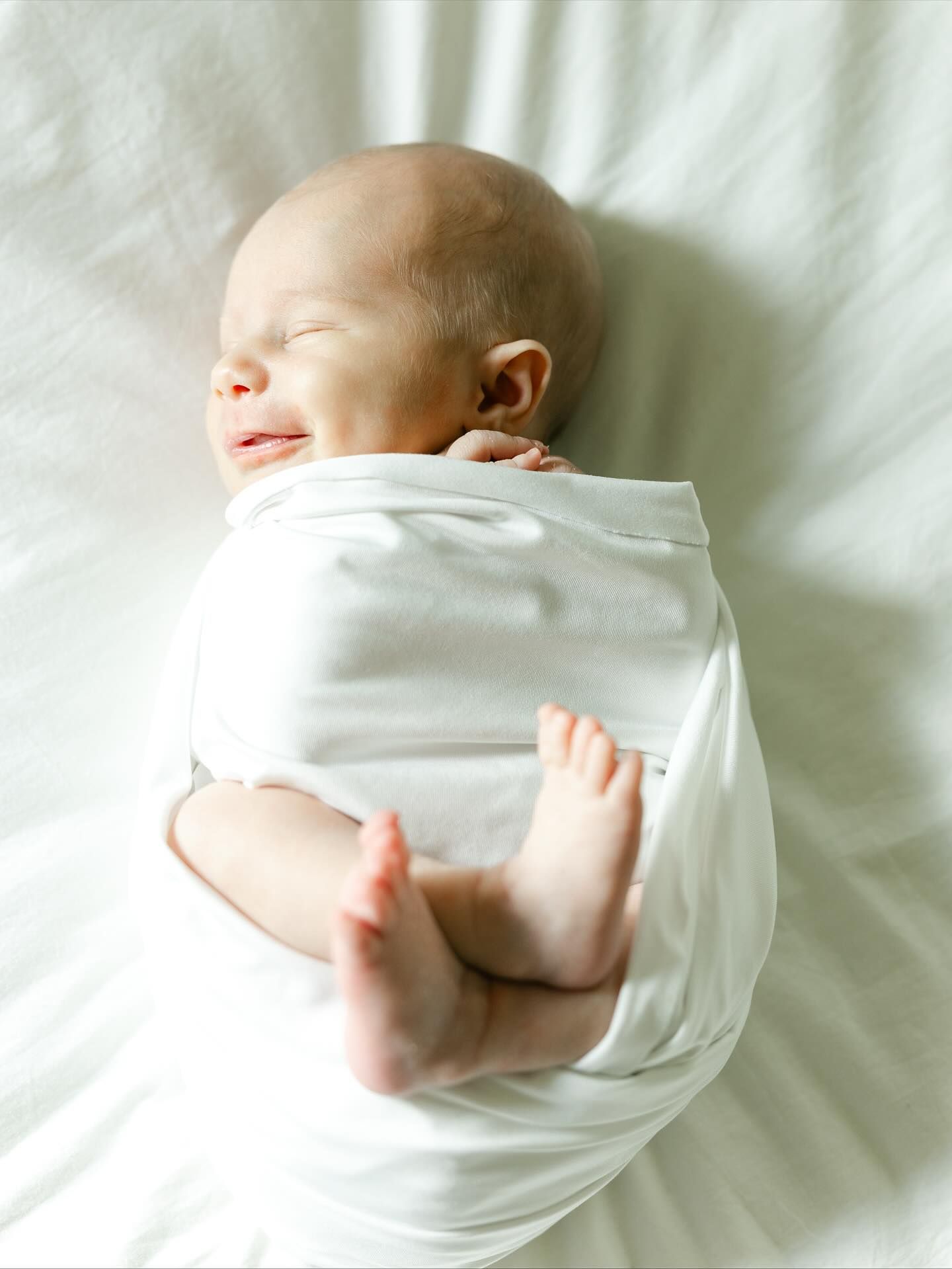 Peaceful sleeping newborn baby swaddled in white cloth with tiny feet peeking out on a soft white surface.