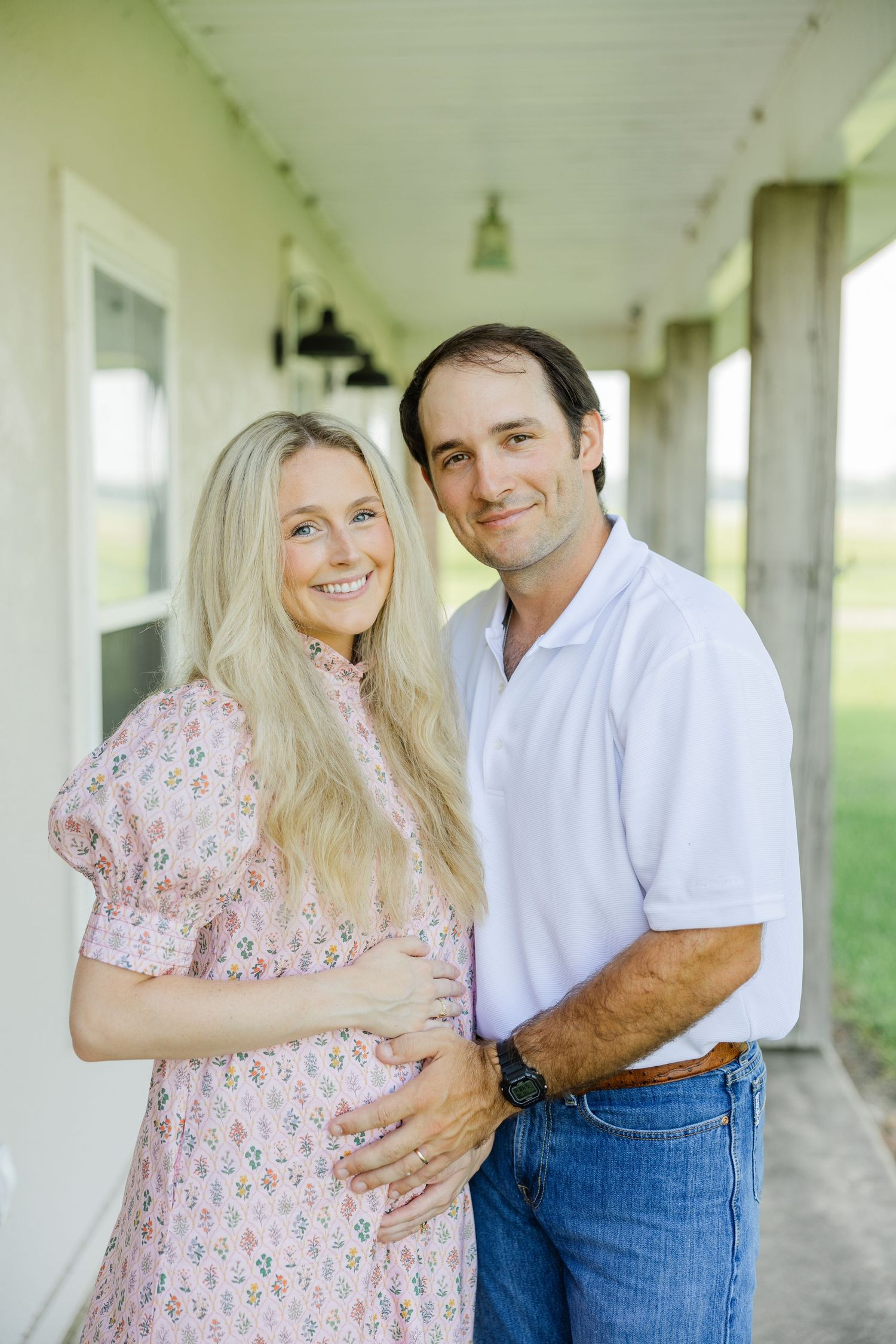 Smiling pregnant woman in pink floral dress with husband on a farmhouse porch.