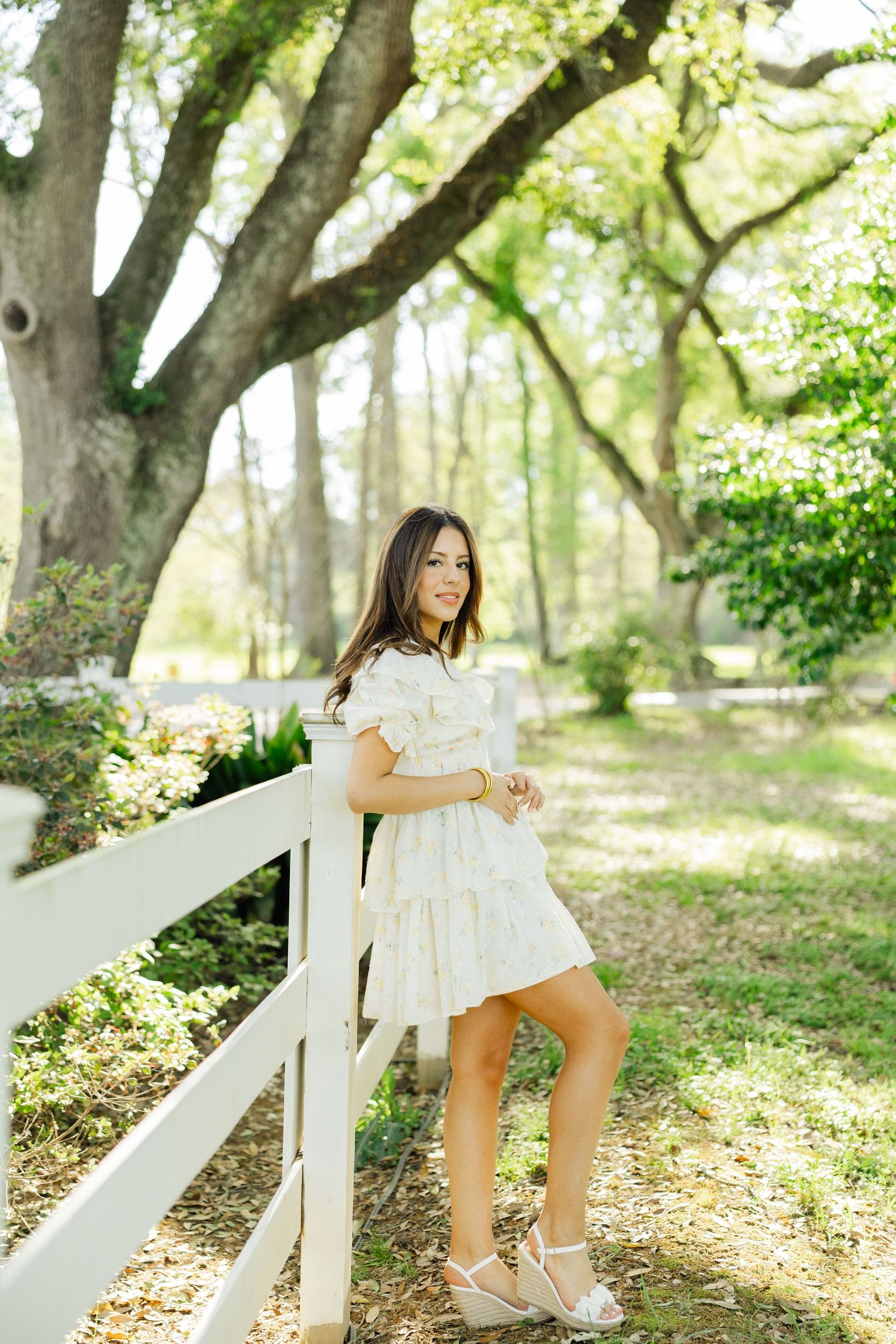 Young woman in white ruffled mini dress leaning on a white fence under large oak trees.