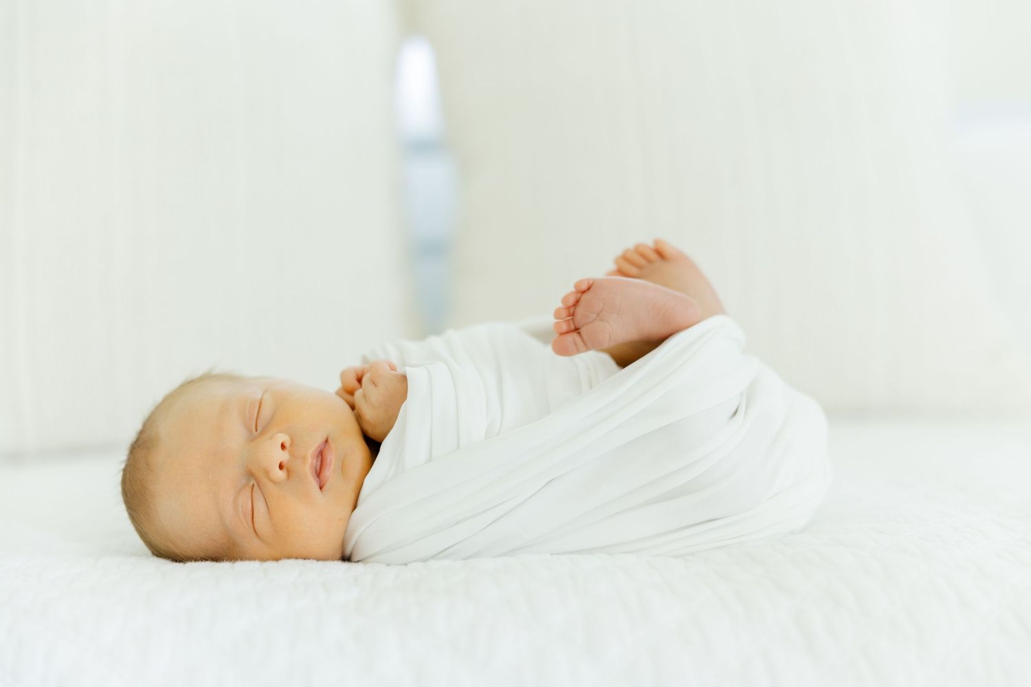 Peaceful newborn baby swaddled in white wrap sleeping on a soft white surface.