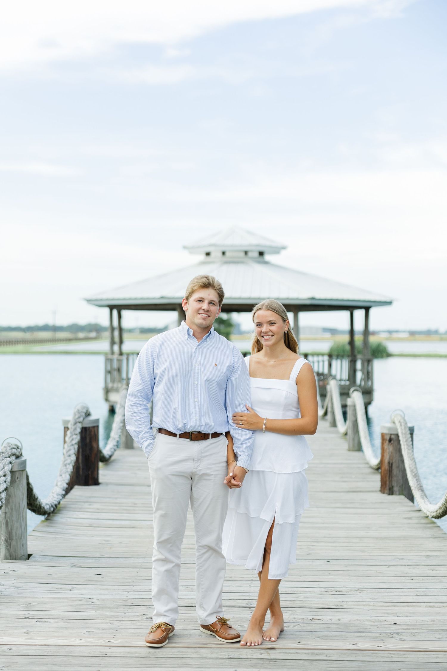Smiling couple holding hands on a wooden dock with a gazebo and open water in the background.