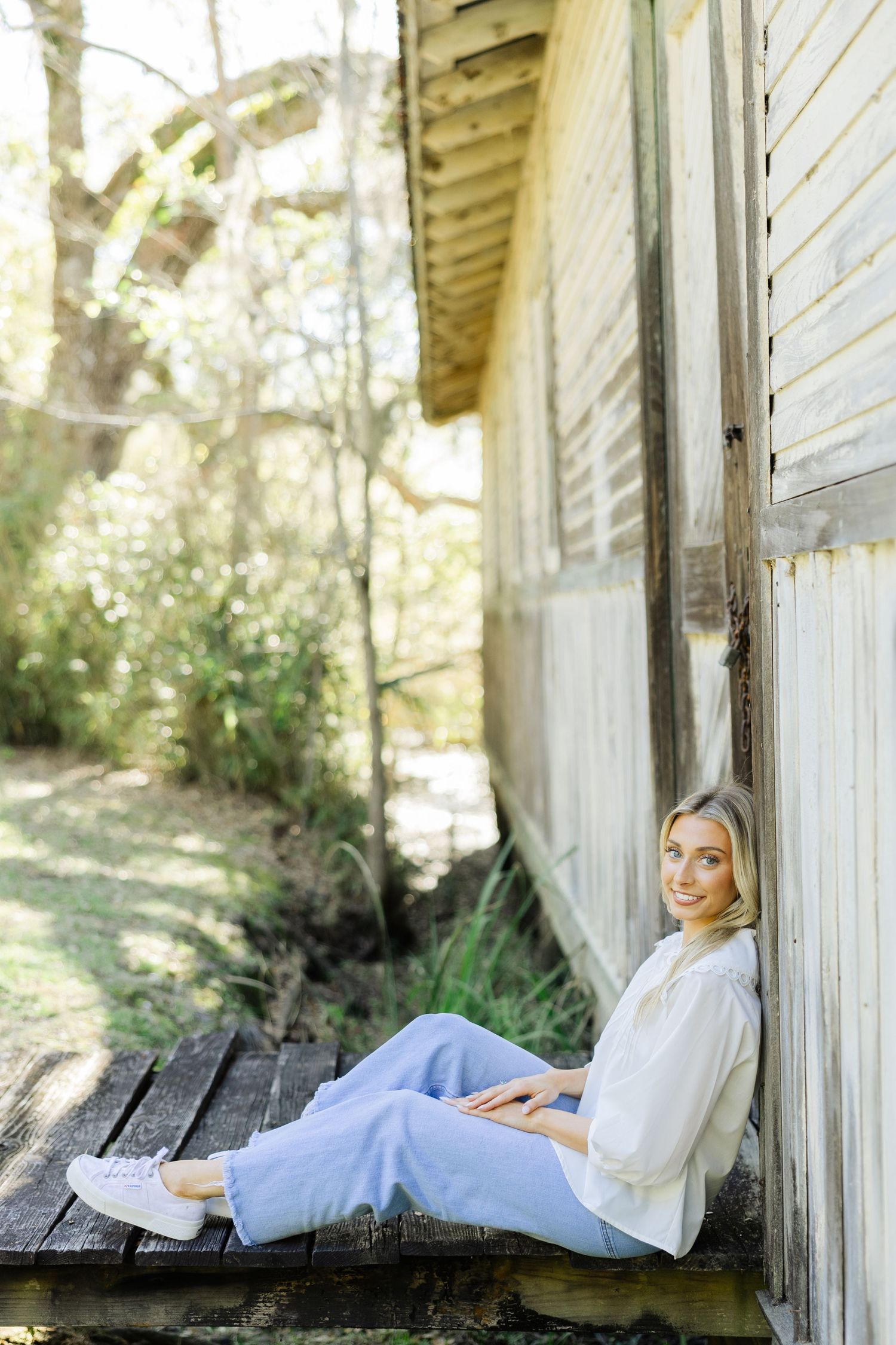 Smiling blonde woman in white blouse and jeans sitting on a rustic wooden porch against a weathered building.
