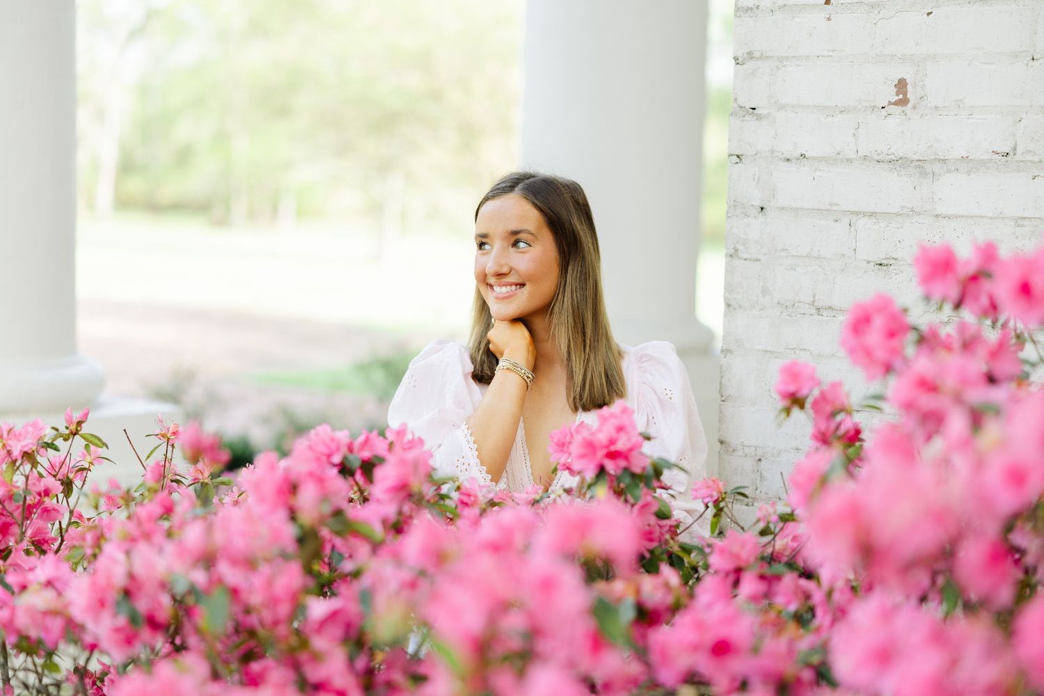 Young woman smiles among vibrant pink azalea blooms beside a white brick column outdoors.