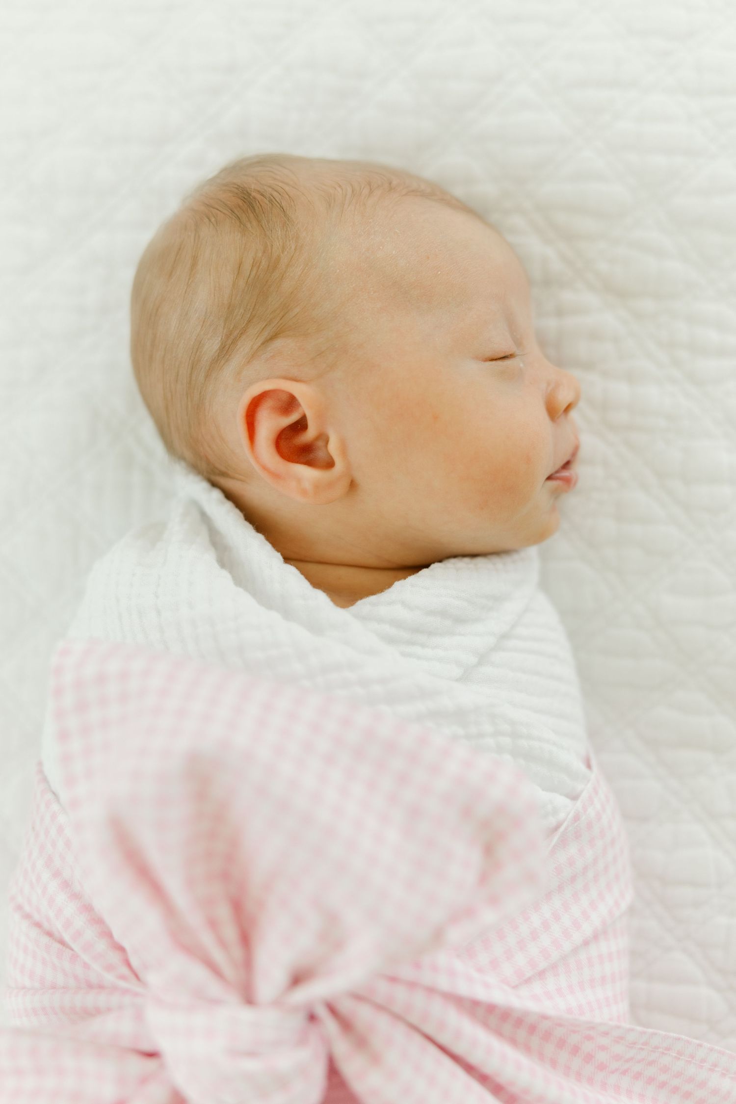 Close-up of a sleeping newborn wrapped in a pink gingham and white swaddle on a quilted white blanket.
