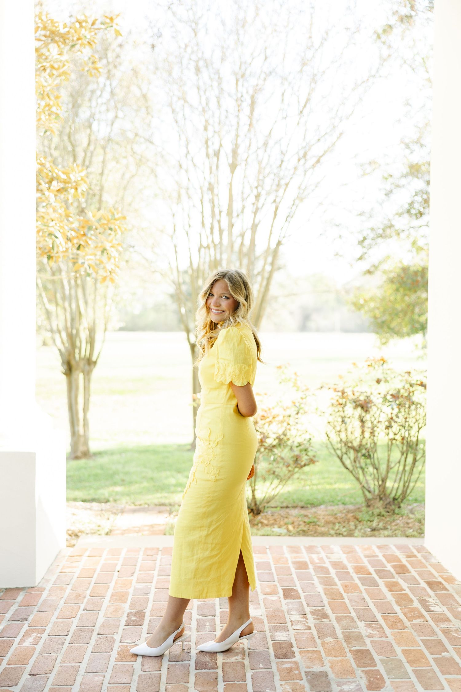 Elegant blonde woman in a yellow midi dress and white heels standing on a sunlit brick porch.