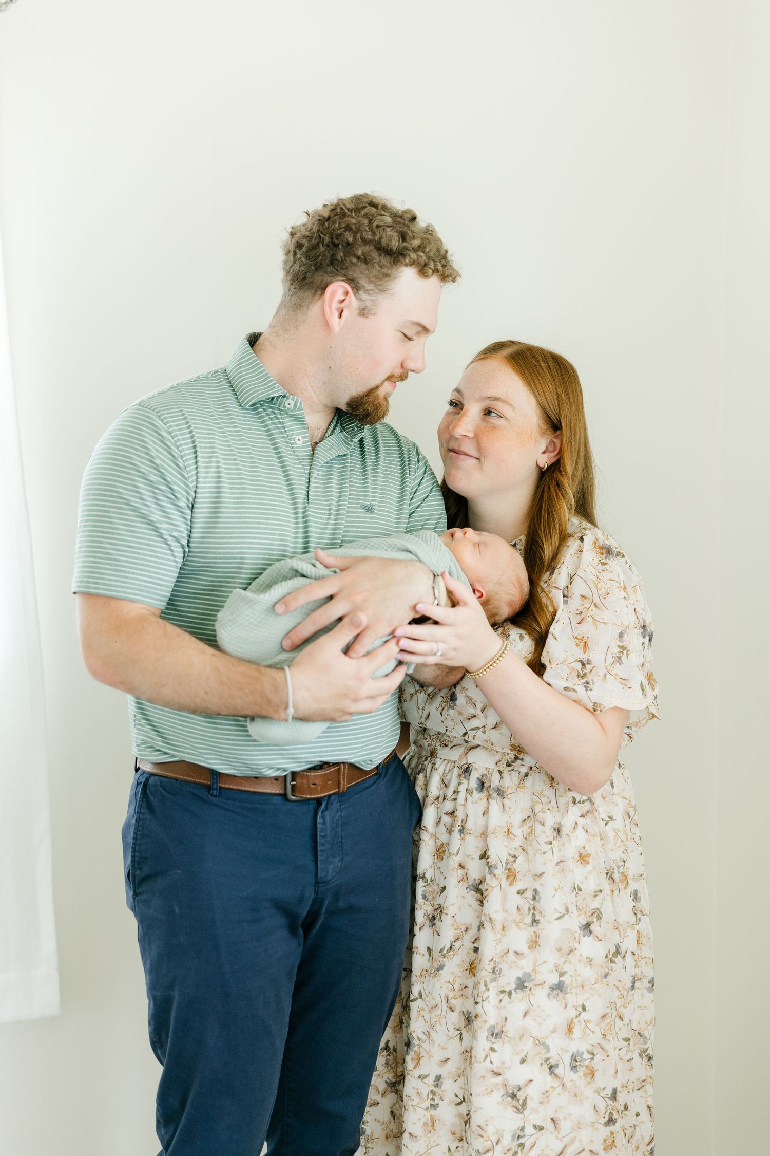 A couple smiles lovingly at their newborn baby, the father in a green polo and mother in a floral dress.