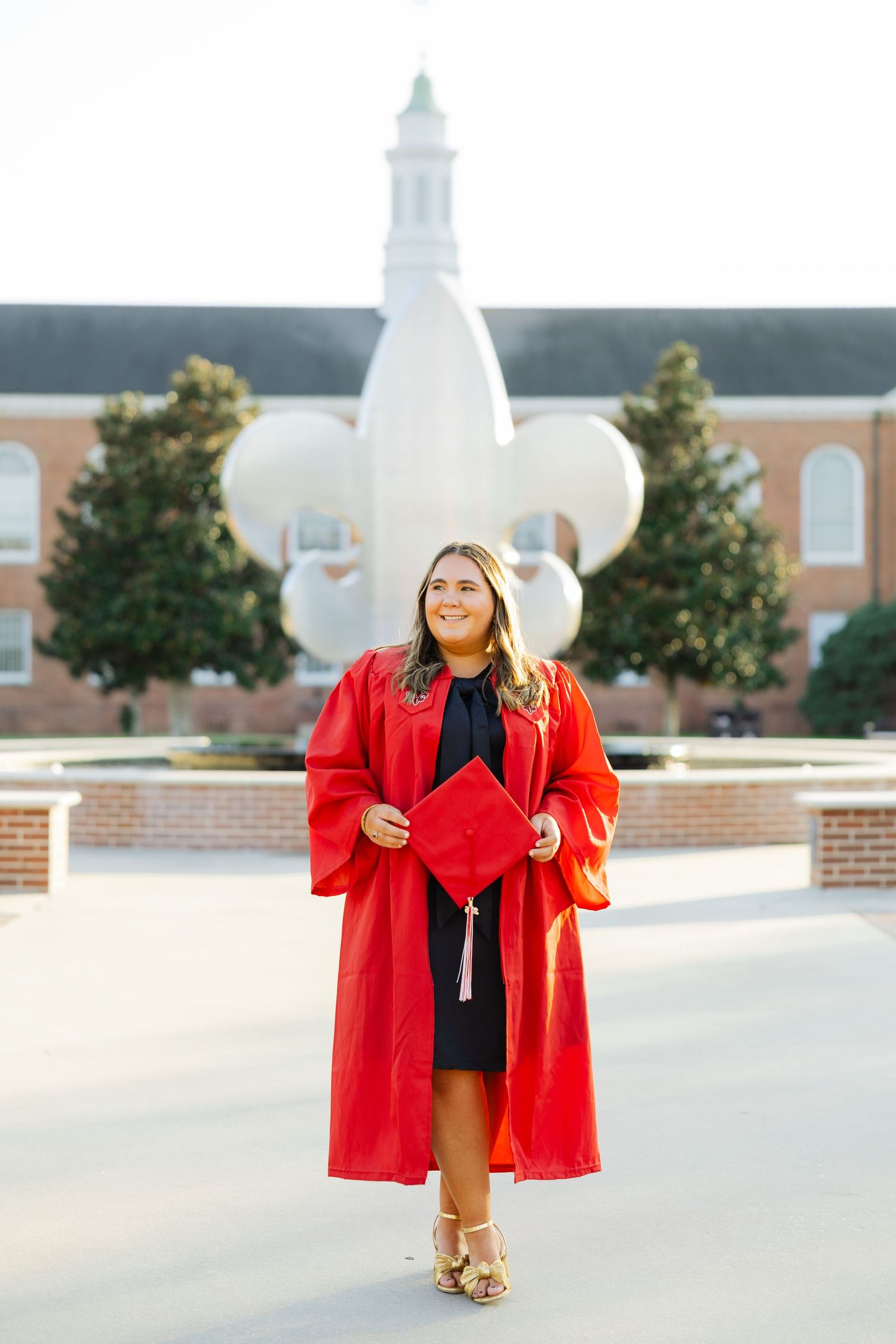 Graduating woman in red cap and gown poses proudly in front of university fountain on campus.