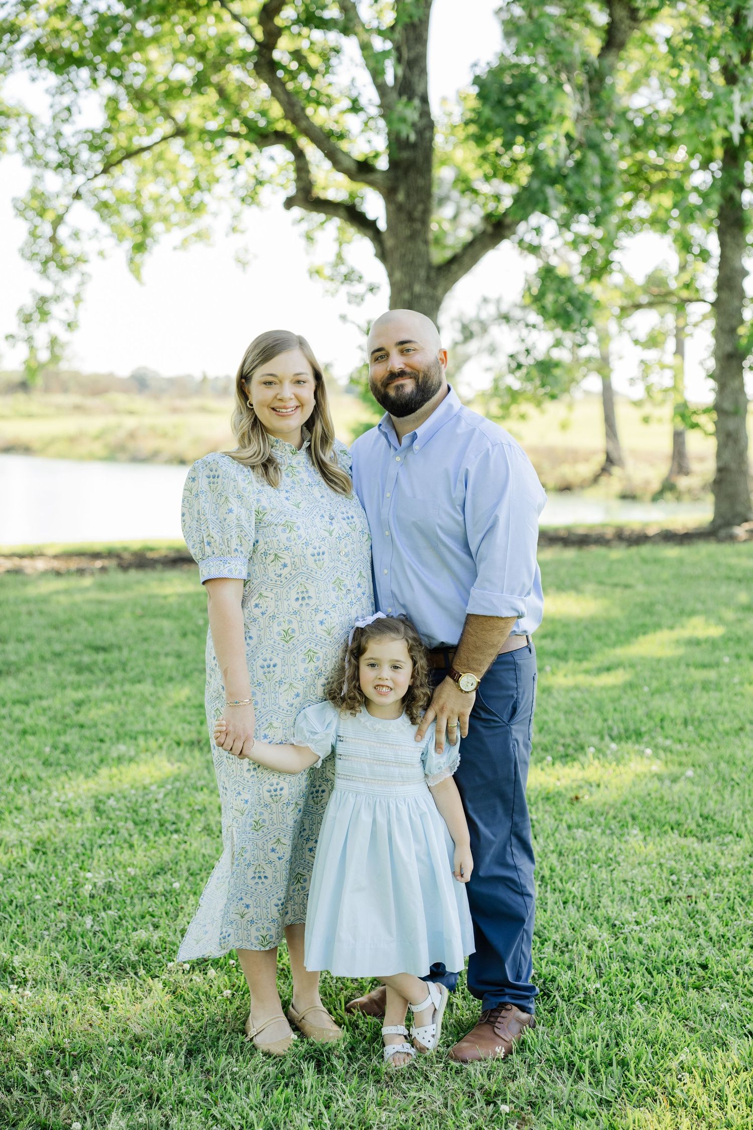 A smiling couple poses outdoors with their young curly-haired daughter in a light blue dress near a pond.