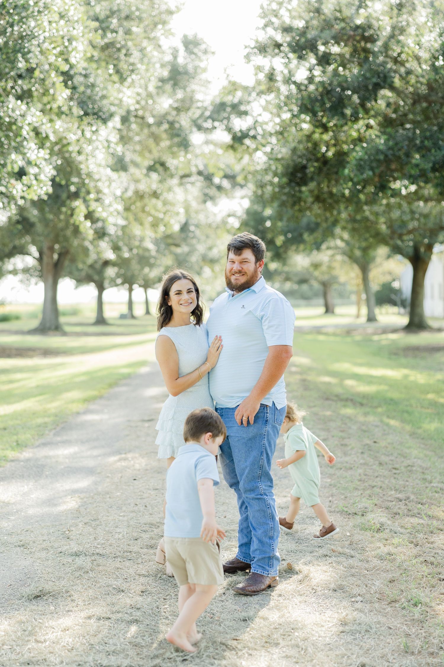 A smiling couple stands on a tree-lined path with two young children playing around them outdoors.