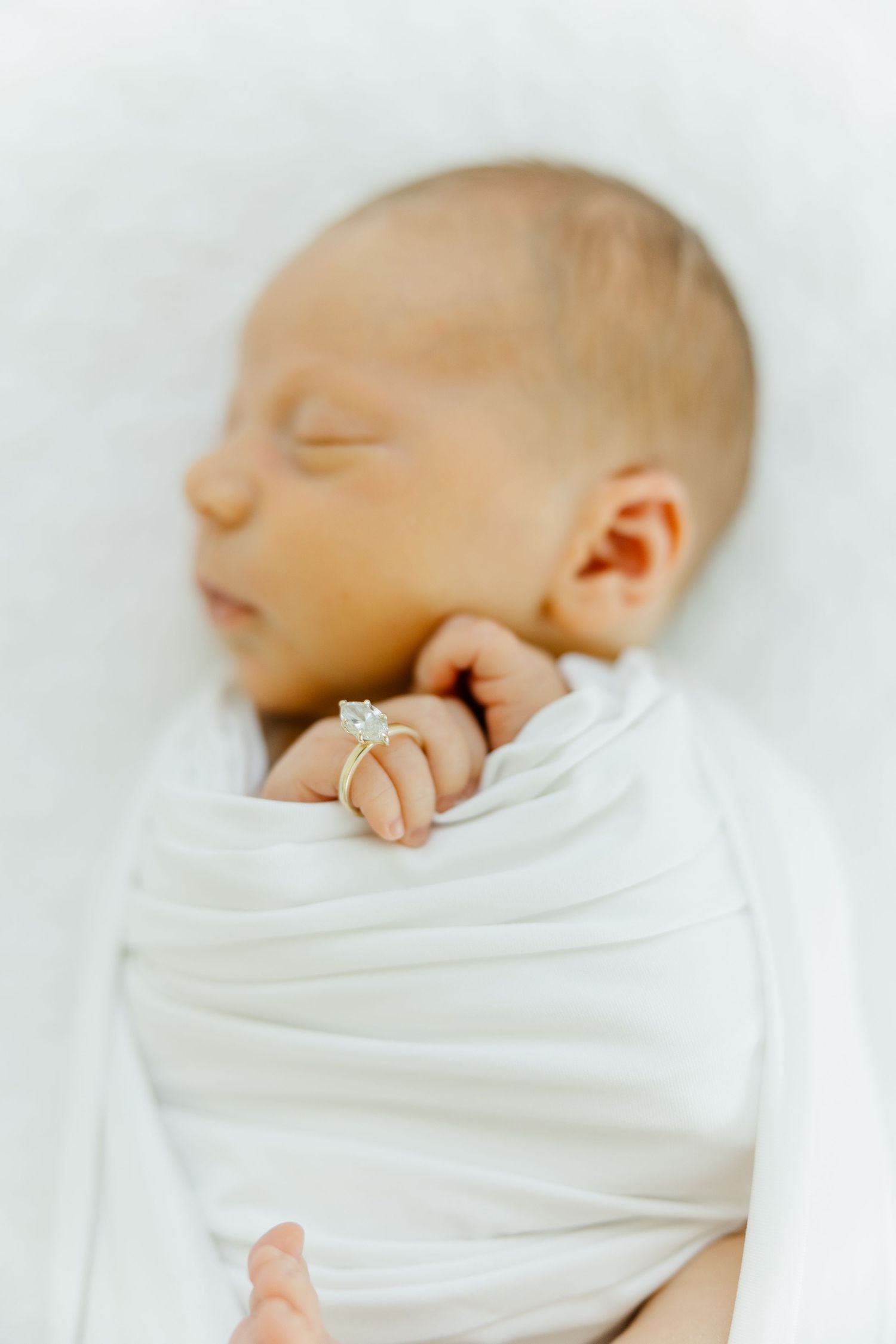 Sleeping swaddled newborn baby with diamond engagement ring placed on tiny fingers.