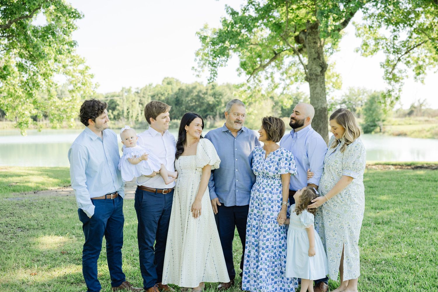 A multigenerational family gathers outdoors under a large tree near a pond for a professional portrait session.
