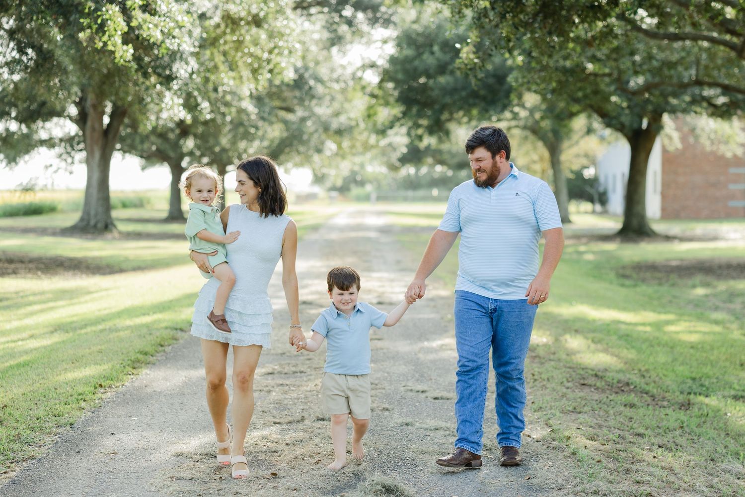 A young family walks hand-in-hand down a sunny dirt path surrounded by lush green trees.