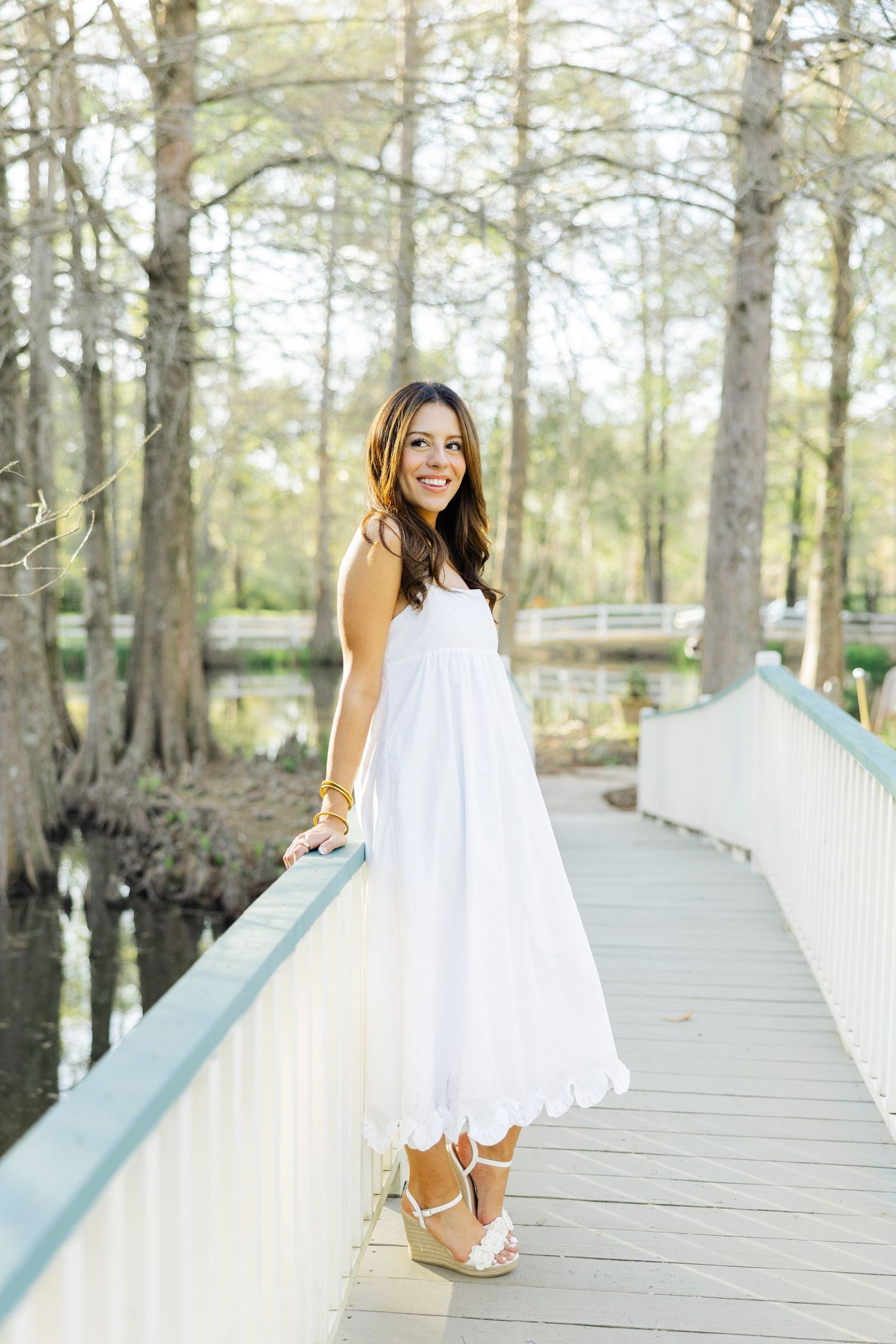 Smiling woman in white eyelet dress and wedge heels poses on a wooden bridge over water.