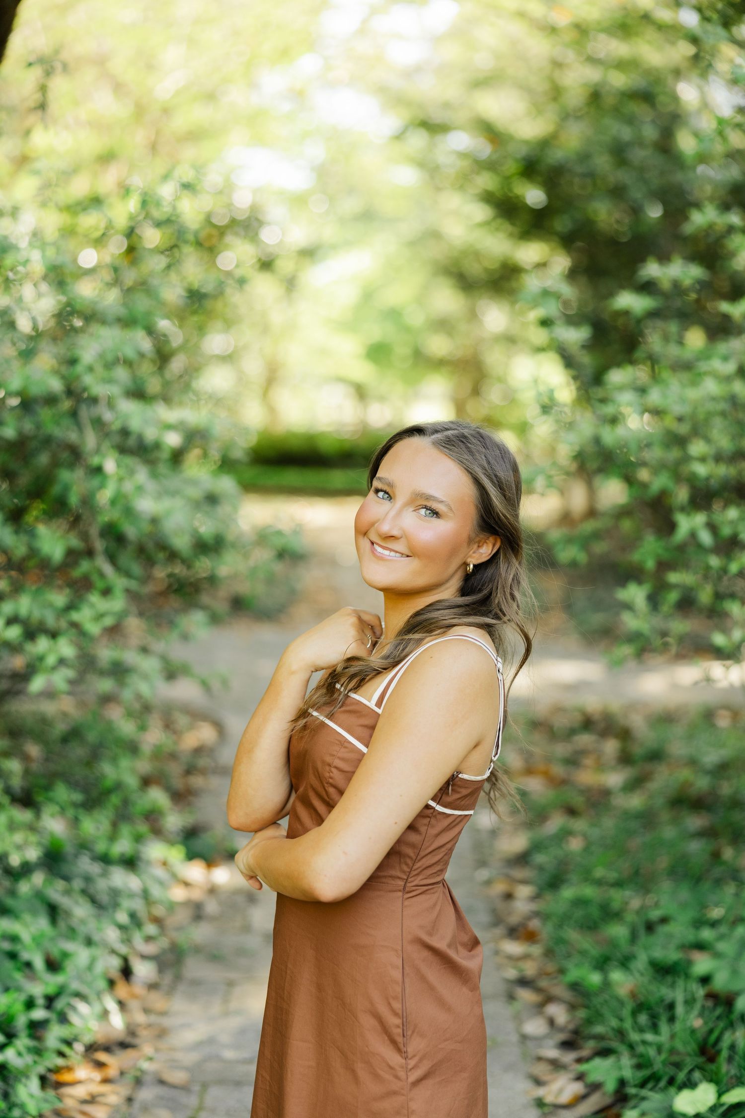 Smiling young woman in brown sundress posing on a tree-lined garden path.