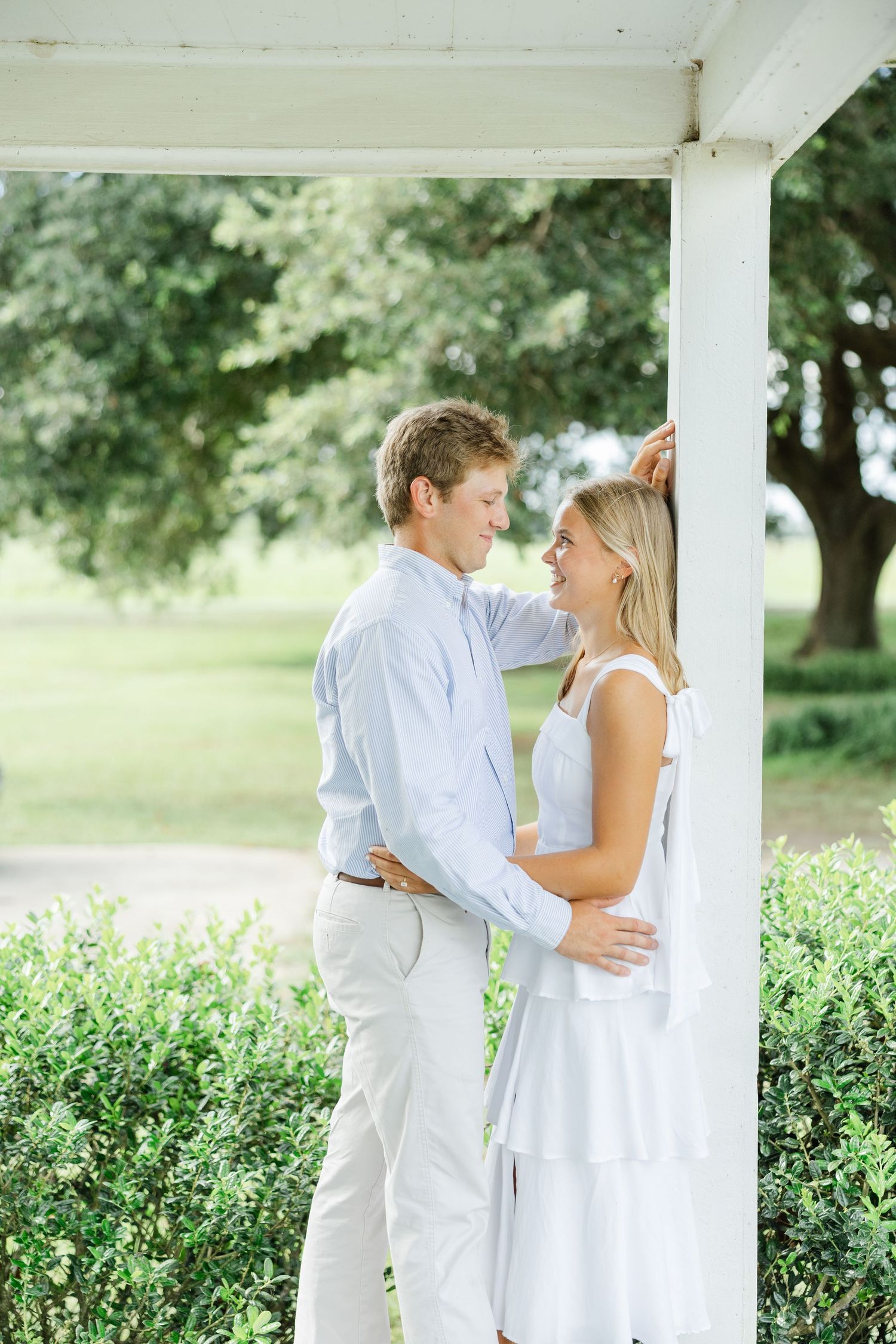 Young couple in white and light blue outfits gazing at each other under a white porch surrounded by greenery.