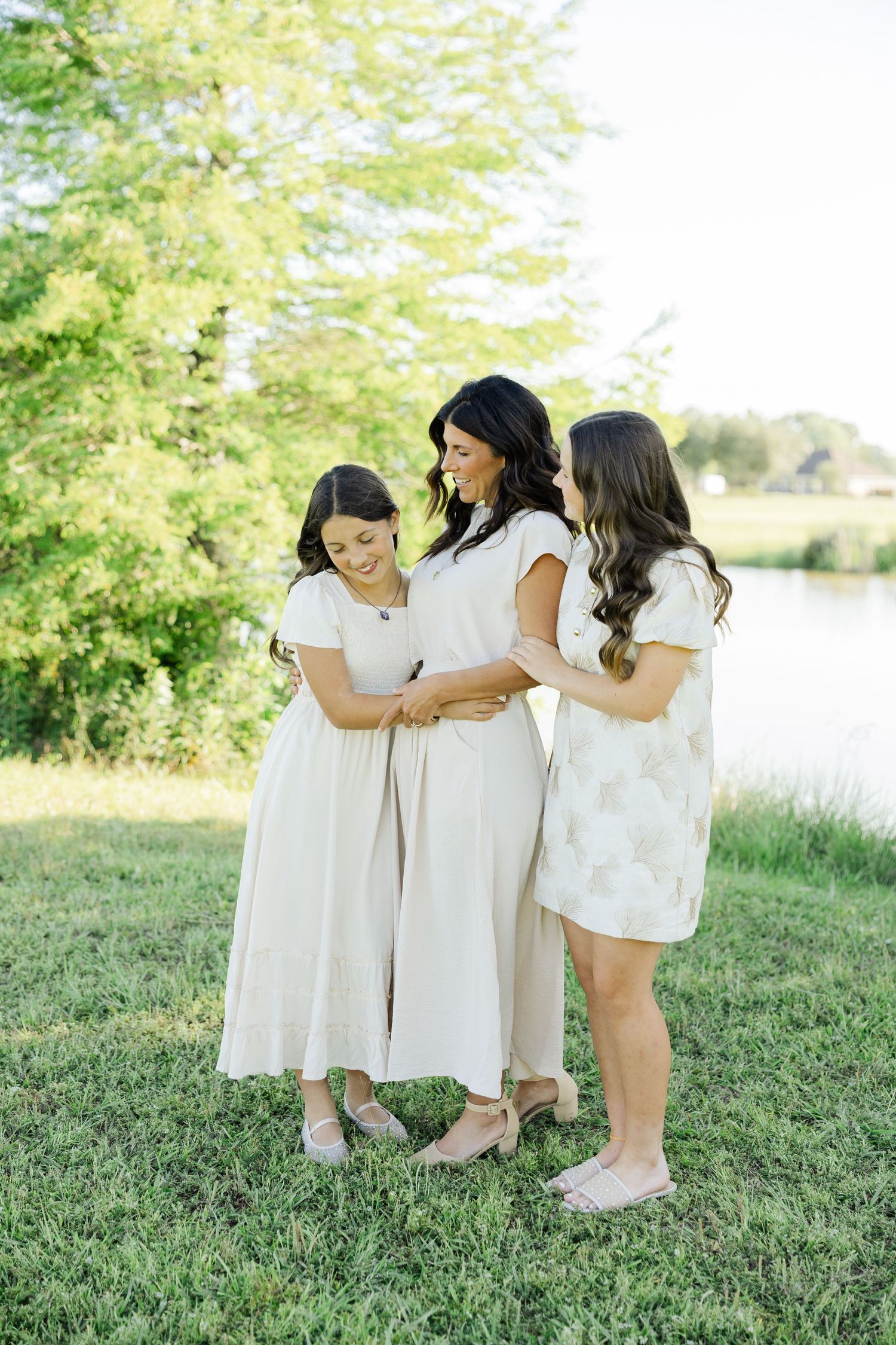 A mother and two daughters share a joyful moment outdoors, wearing coordinating neutral dresses near a serene pond.
