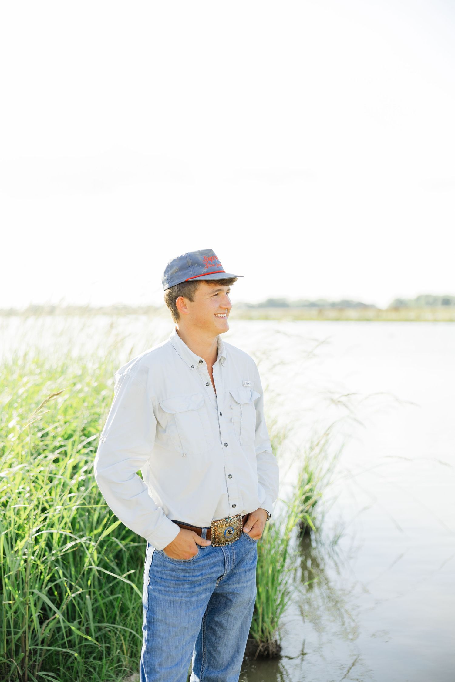 Smiling young man in white shirt and cap stands by tall marsh grass near a lake.
