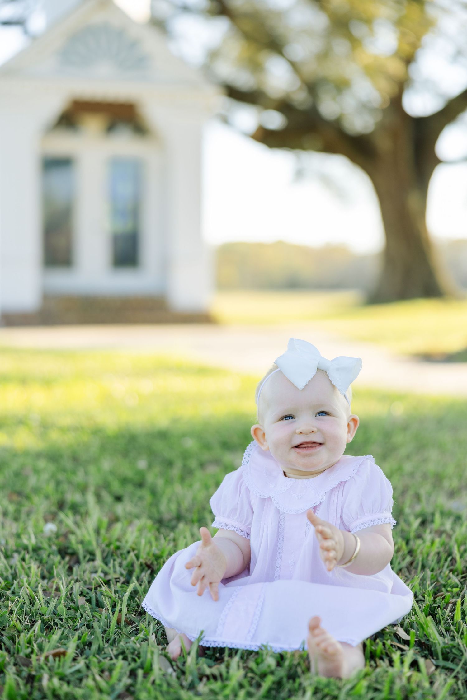 Happy baby girl in pink dress and white bow sitting on green grass clapping hands.