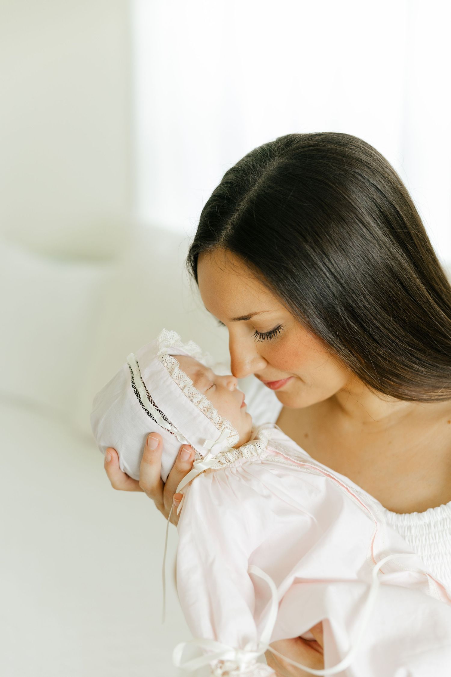 Dark-haired mother gently nuzzling her newborn baby in a white bonnet in a bright room.