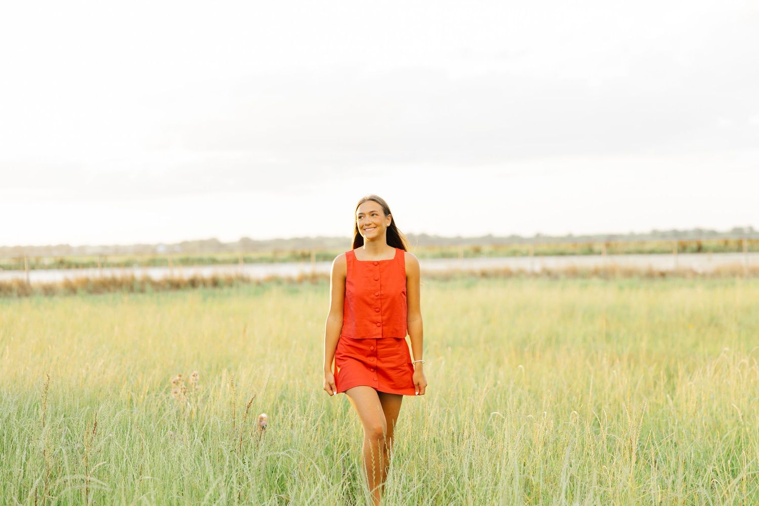 Young woman in red dress walking through tall green grass field near water.