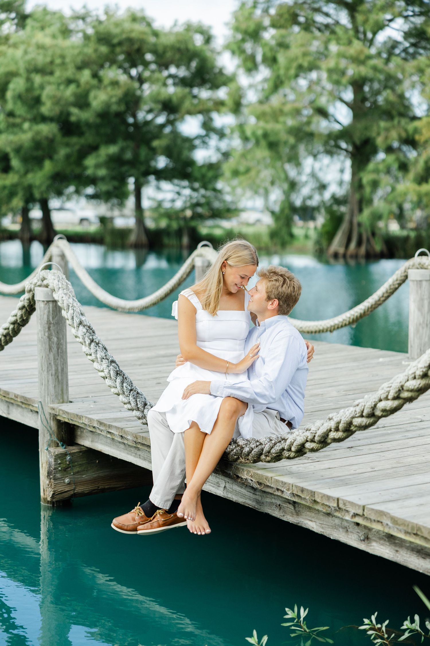 Romantic couple sitting close together on a wooden dock over turquoise water surrounded by trees.