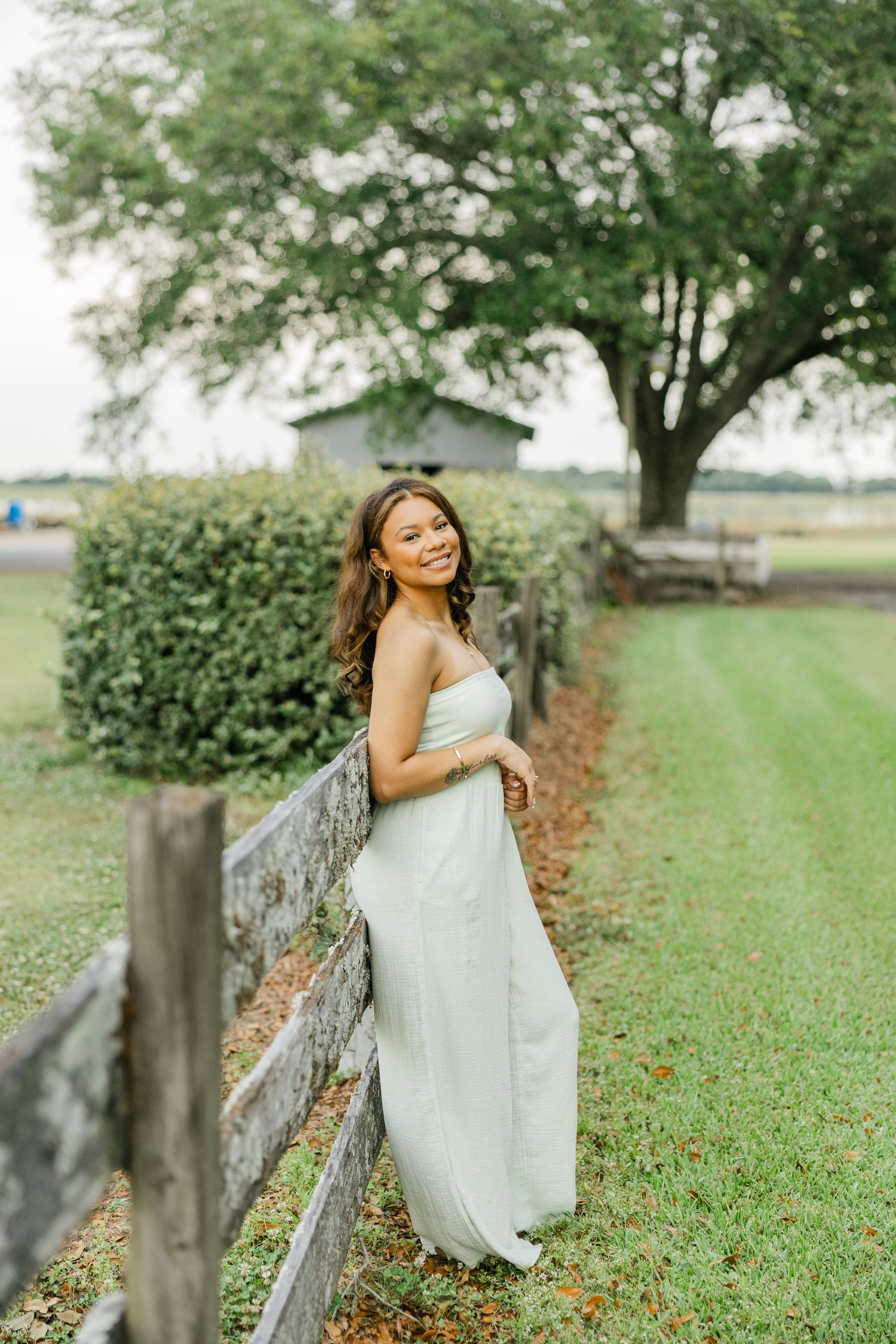 Elegant young woman in mint green maxi dress posing by a wooden fence with trees in background.