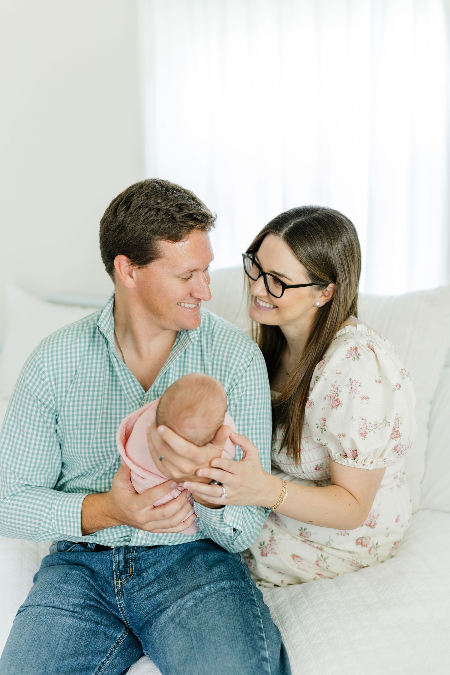 Happy parents in a bright room smiling down at their newborn baby wrapped in a pink swaddle.