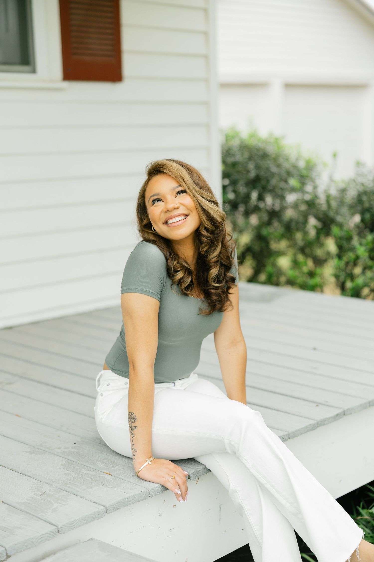 Smiling woman in a sage green top and white pants sitting on a porch step looking upward joyfully.