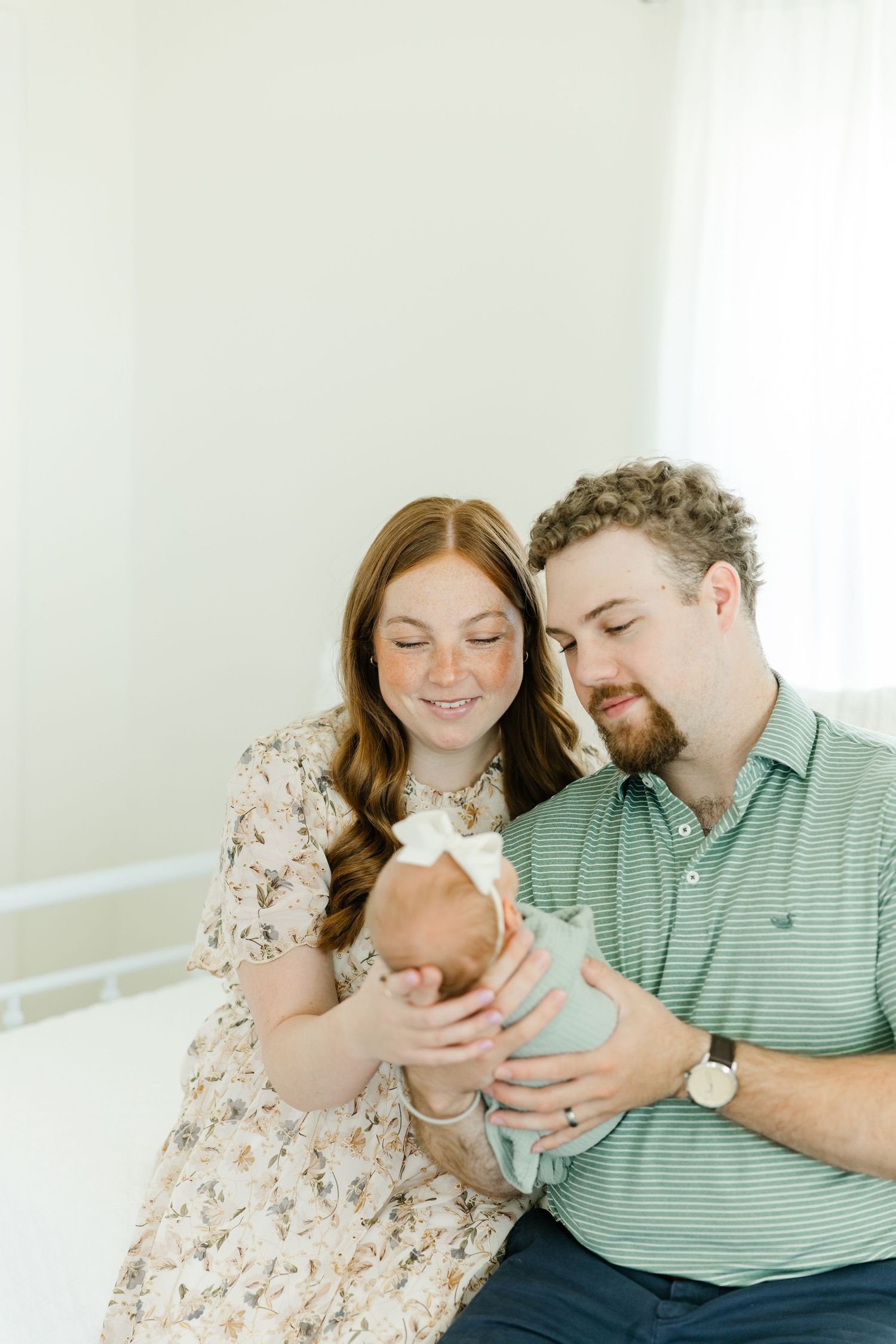 Smiling red-haired mother and bearded father in green polo gently hold their newborn baby girl with bow.