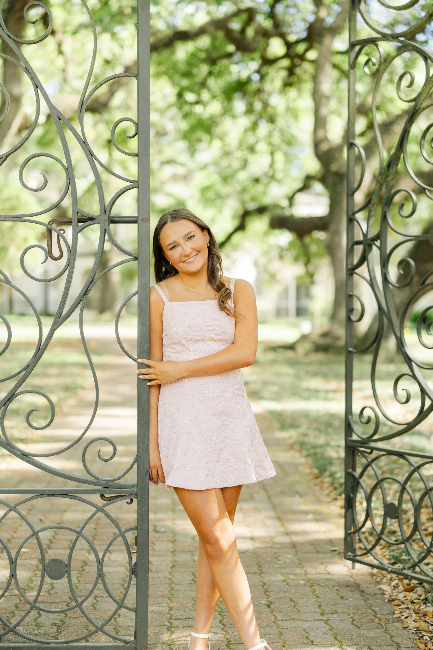 Smiling young woman in light pink mini dress poses in ornate wrought iron garden gate.