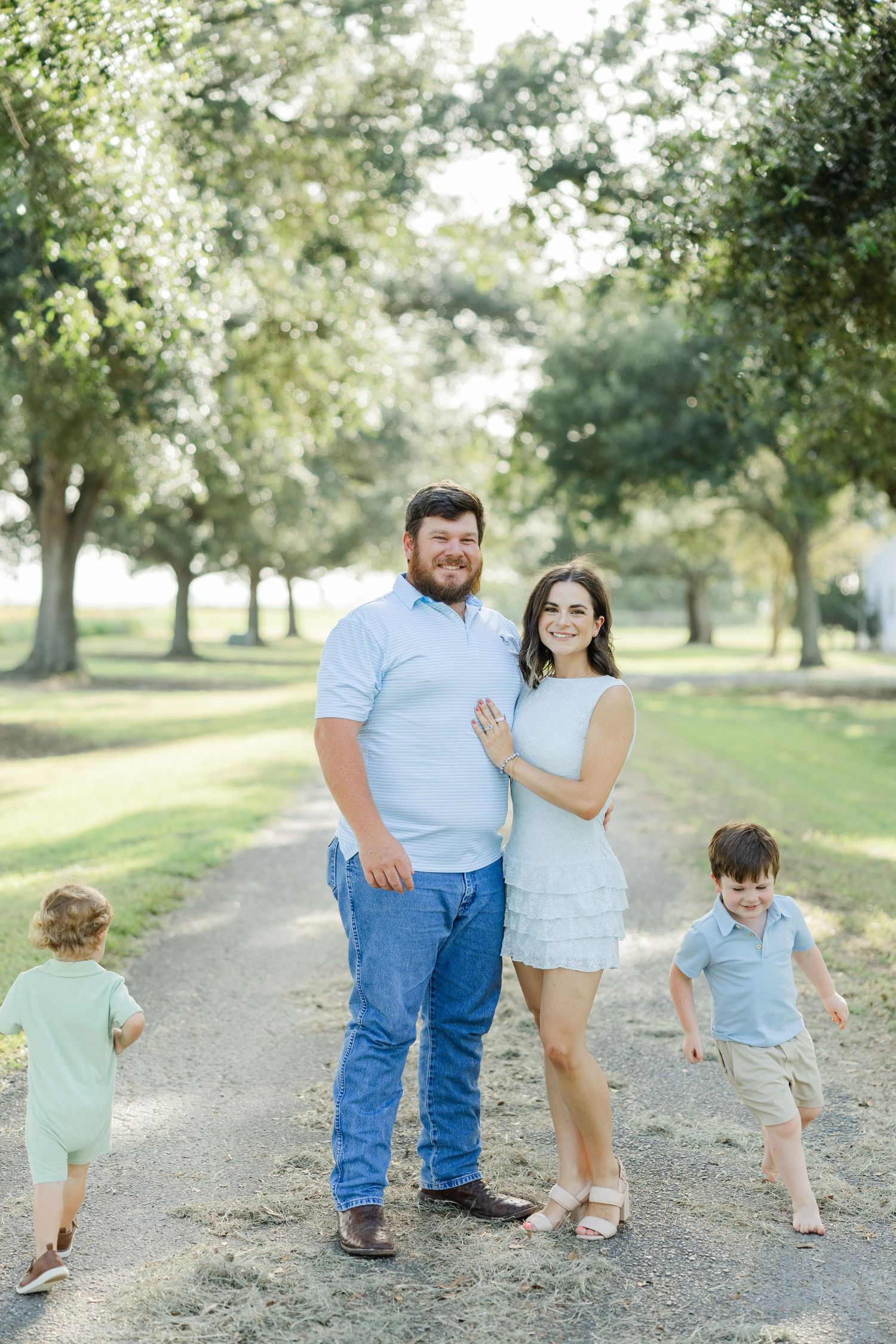 Happy family of four posing on a tree-lined dirt path on a sunny day in casual blue outfits.