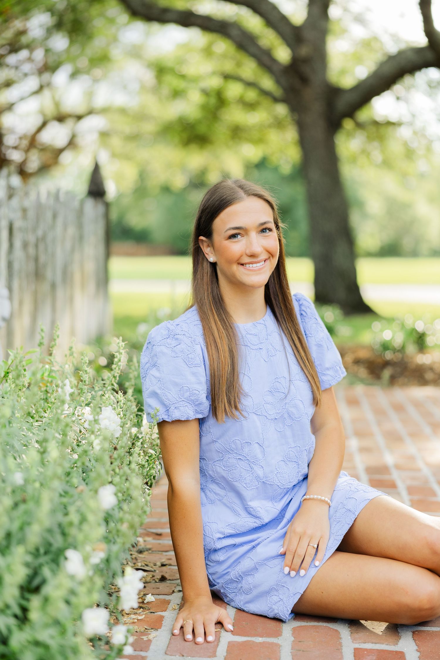 Smiling brunette in blue puff-sleeve dress sitting on brick path beside white flowers.