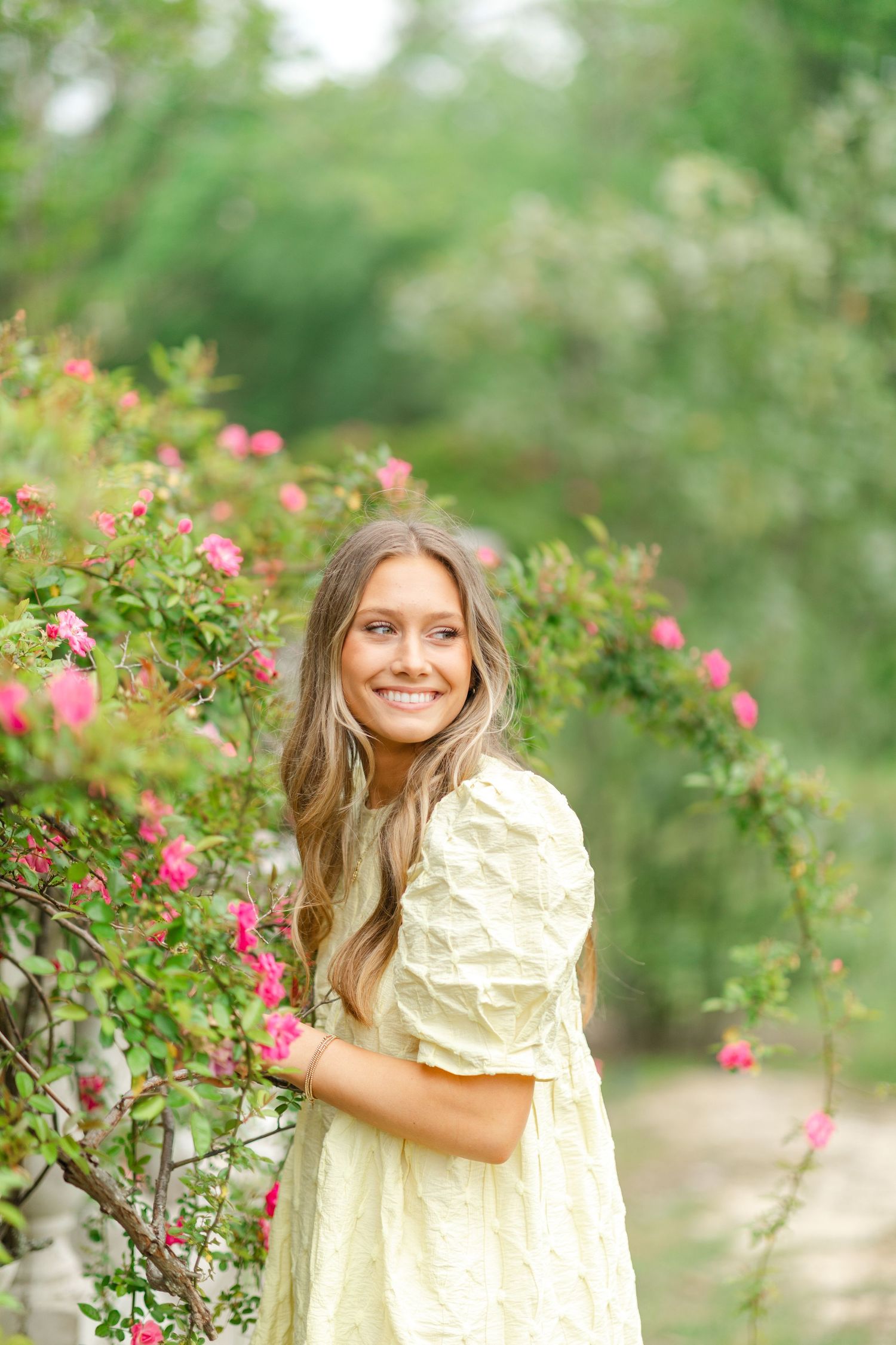 Smiling teenage girl in cream dress standing beside blooming pink rose bushes in a lush garden.