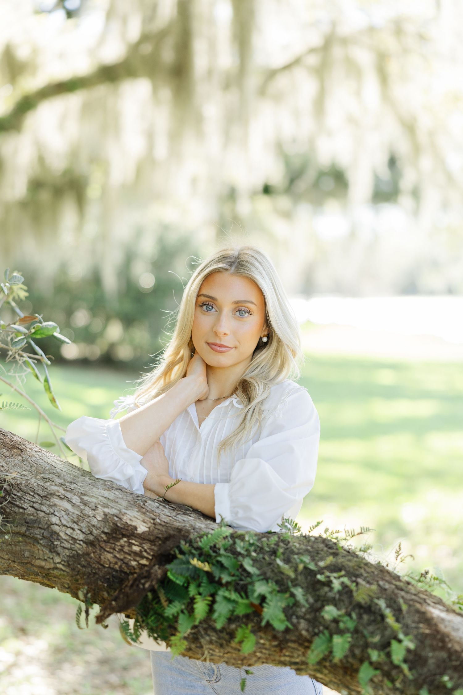 Blonde woman in a white shirt leaning on a moss-covered tree branch in a sunlit park setting.
