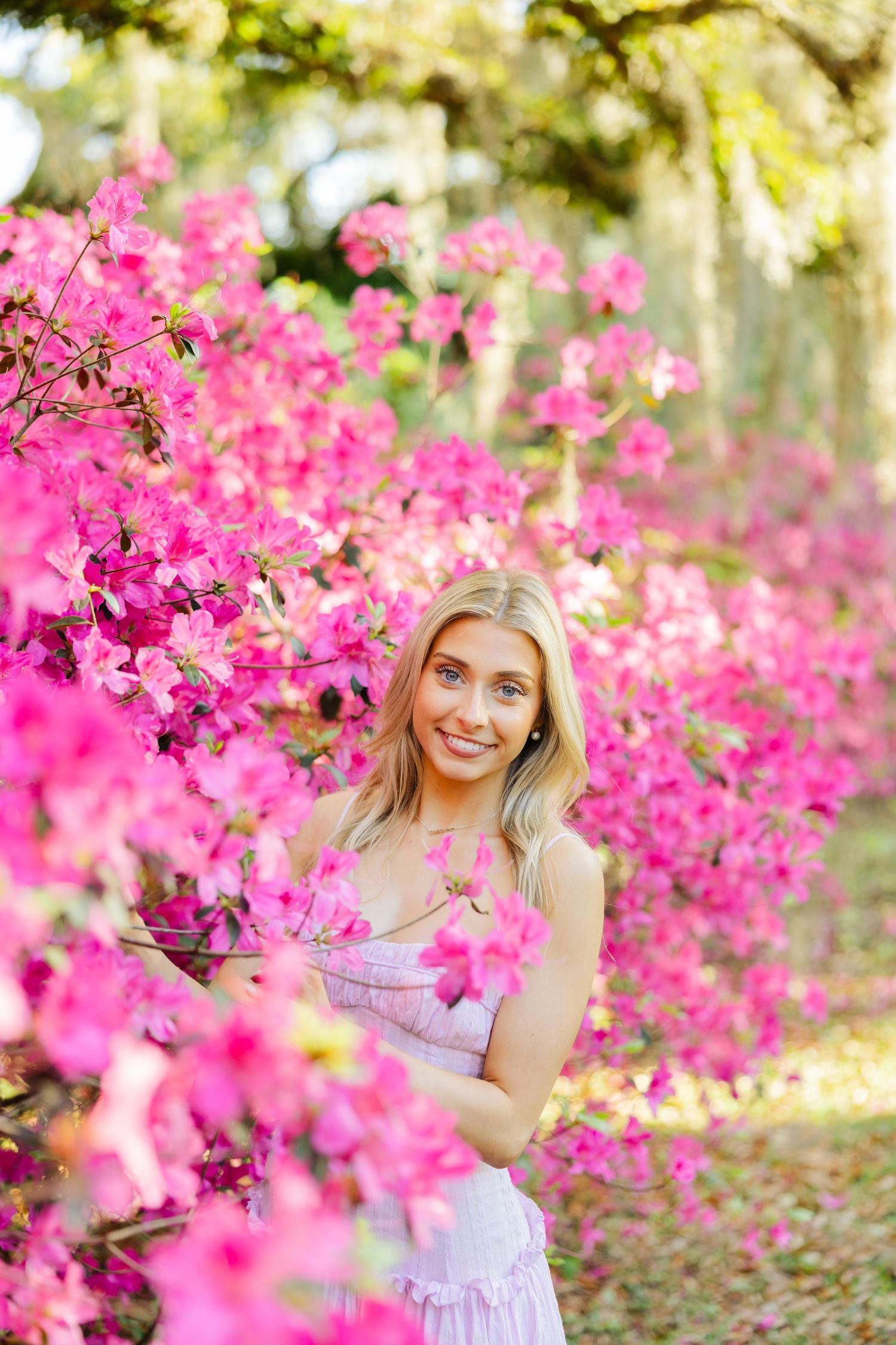 Smiling blonde woman in lavender dress poses among vibrant pink azalea blooms in a garden.