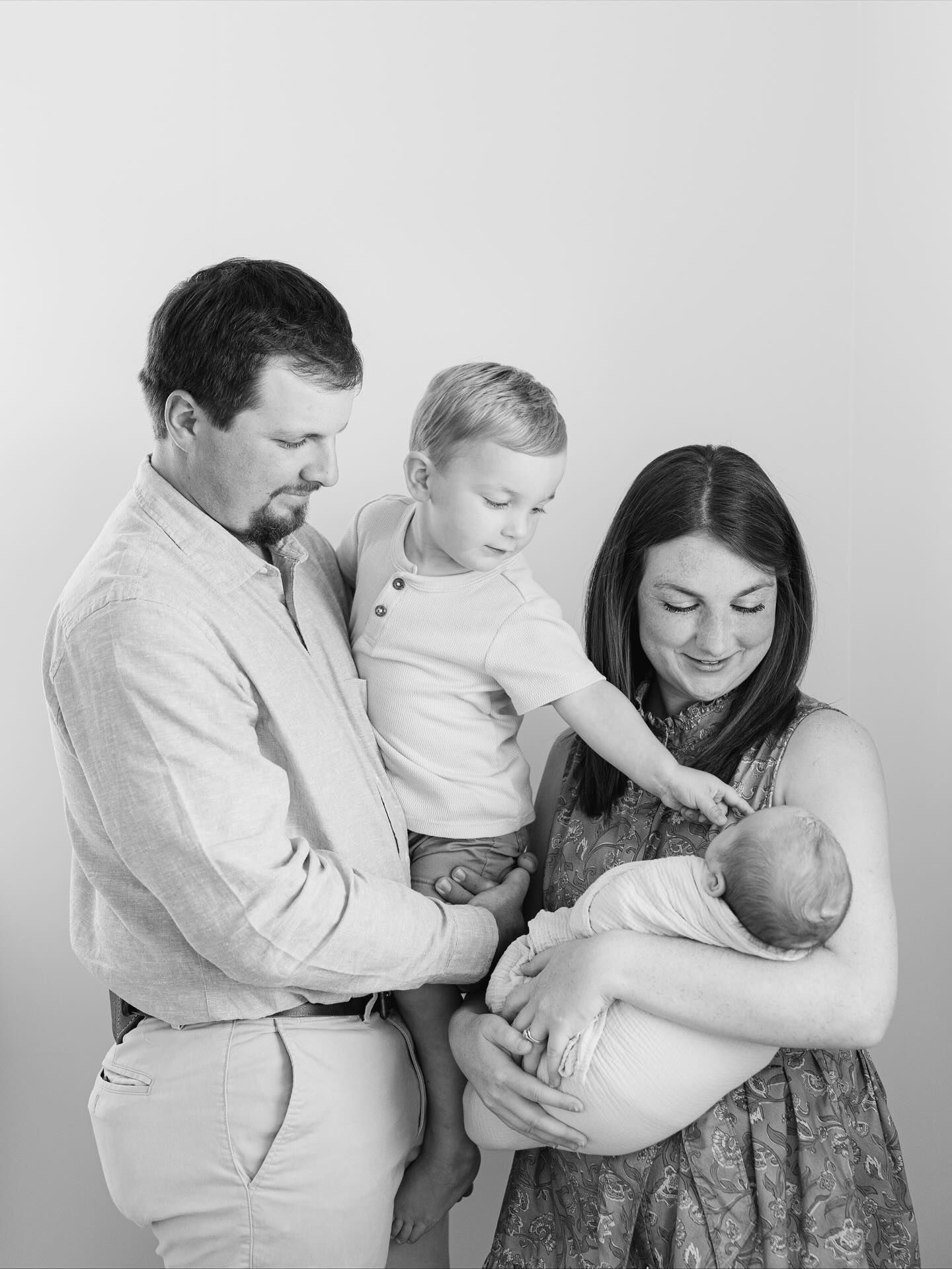 Black and white family portrait of parents with toddler son and newborn baby in a bright minimalist studio setting.
