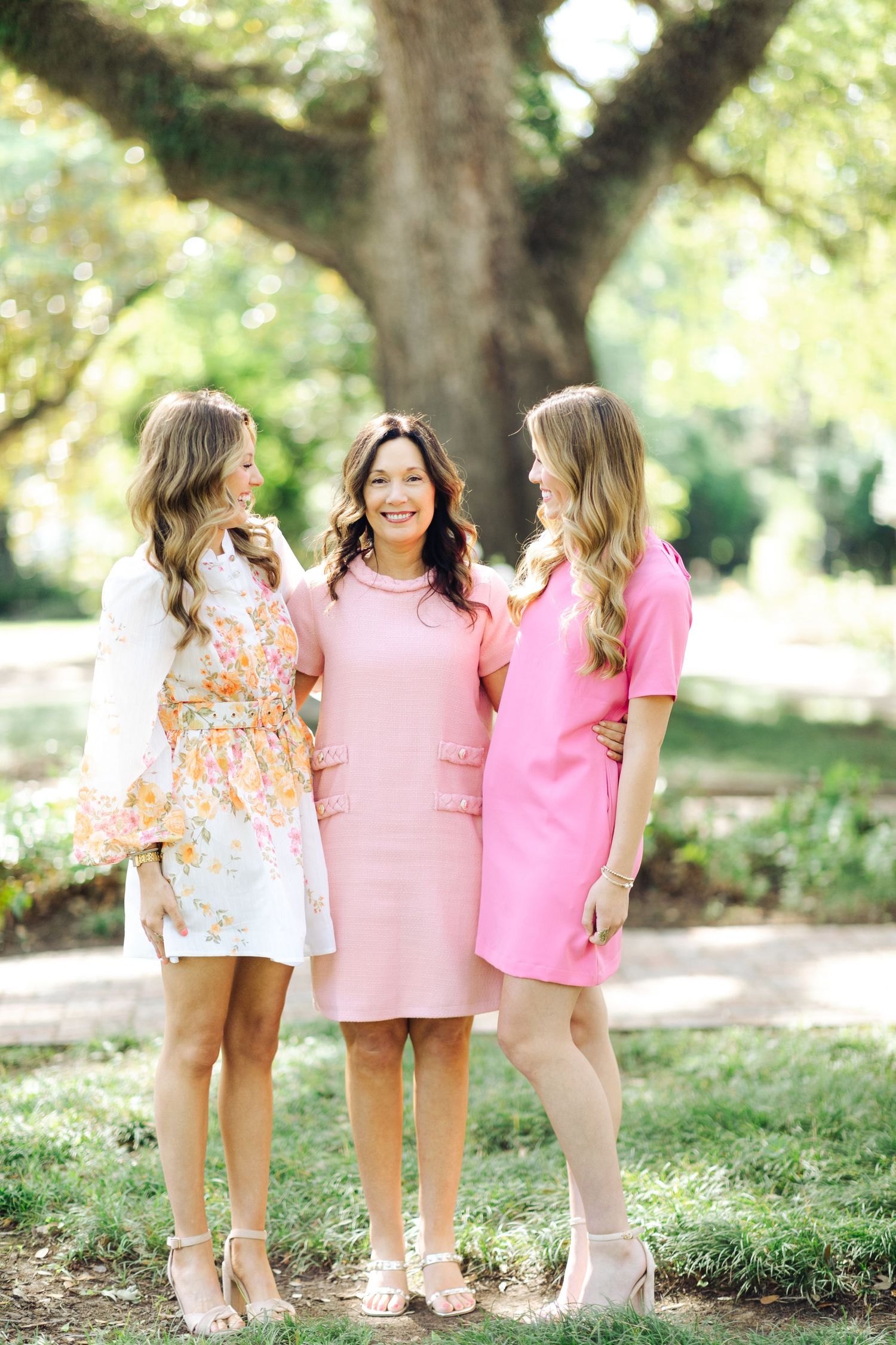 Three women in pink and floral dresses smiling together outdoors under a large tree during a family photo session.
