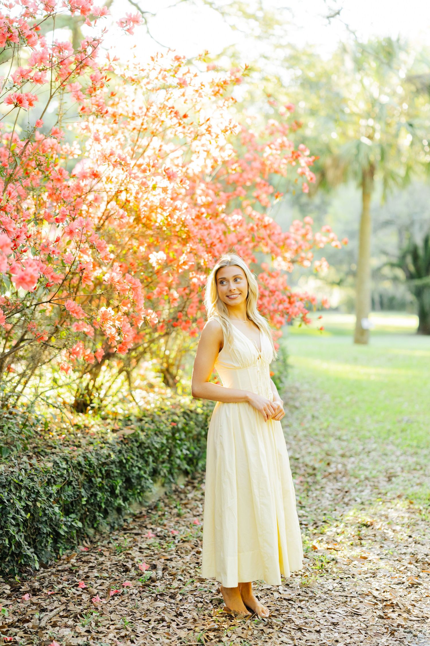 Blonde woman in a yellow midi dress standing on a leaf-covered path beside vibrant pink flowering trees.