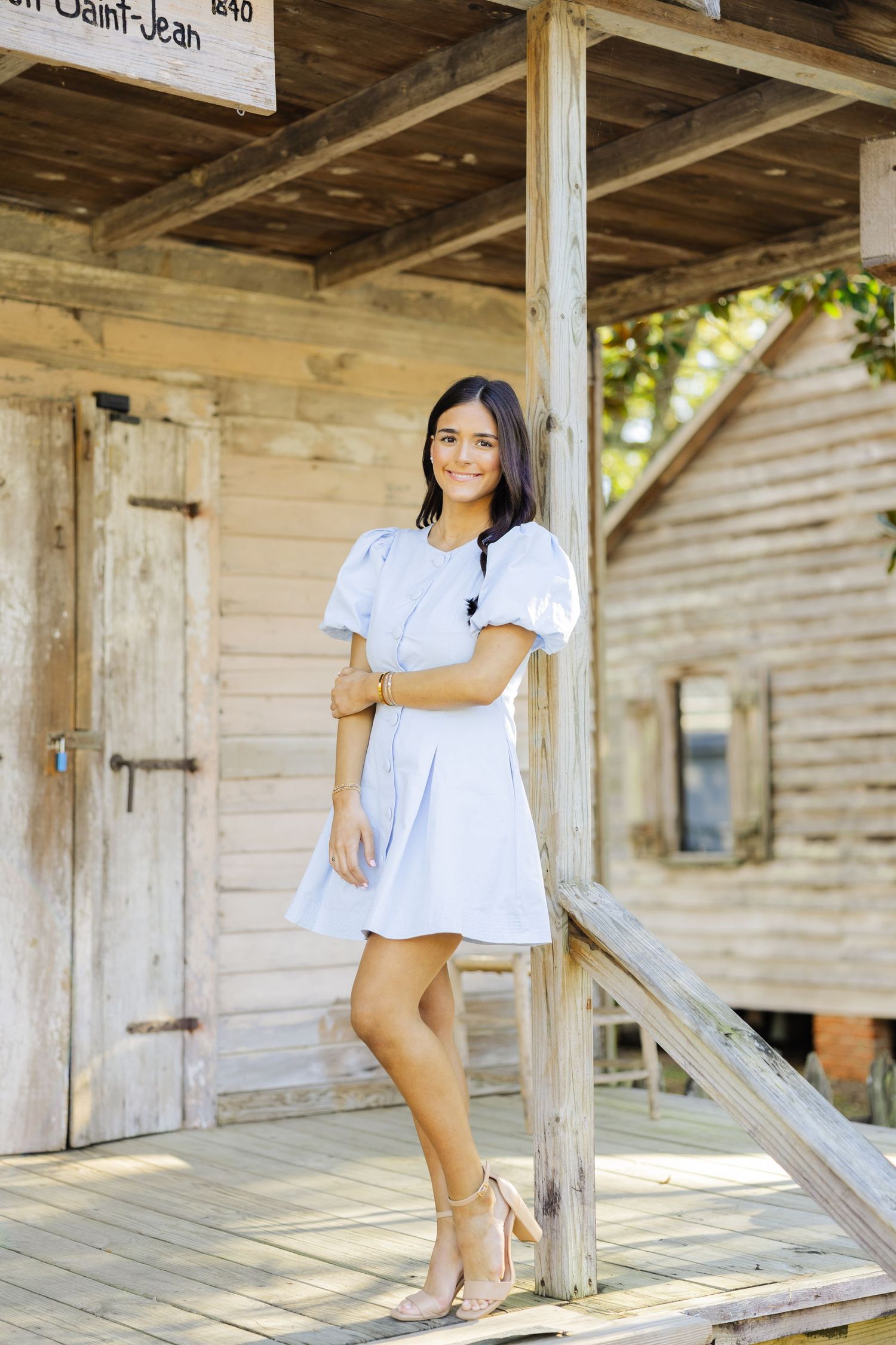 Smiling young woman in light blue puff-sleeve dress leaning on a wooden post of a rustic building.