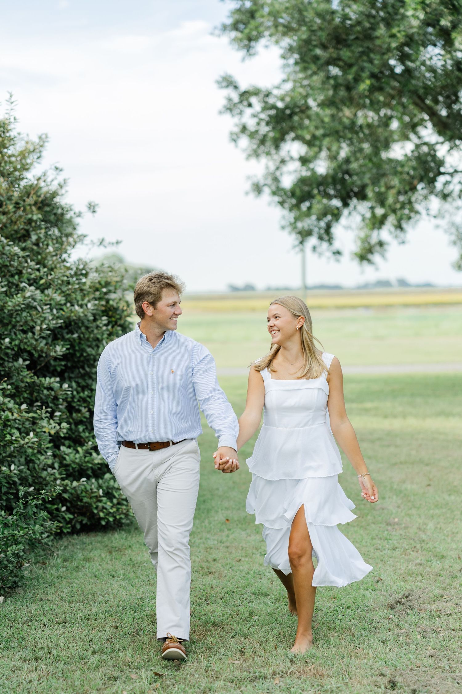 Couple holding hands and smiling while walking across a green lawn with trees and open fields behind them.
