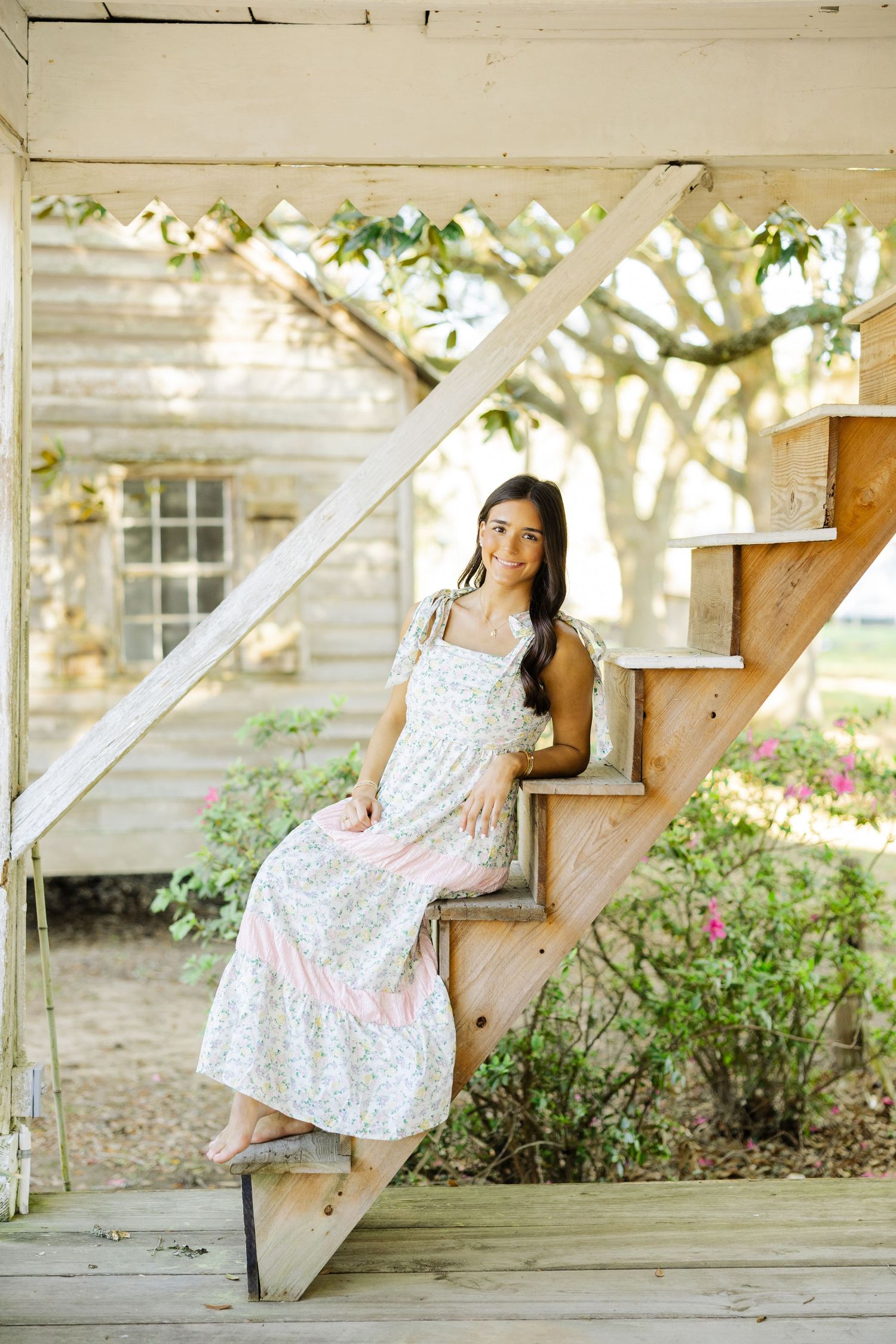 Young woman in floral maxi dress sitting on wooden stairs outside a rustic white farmhouse.