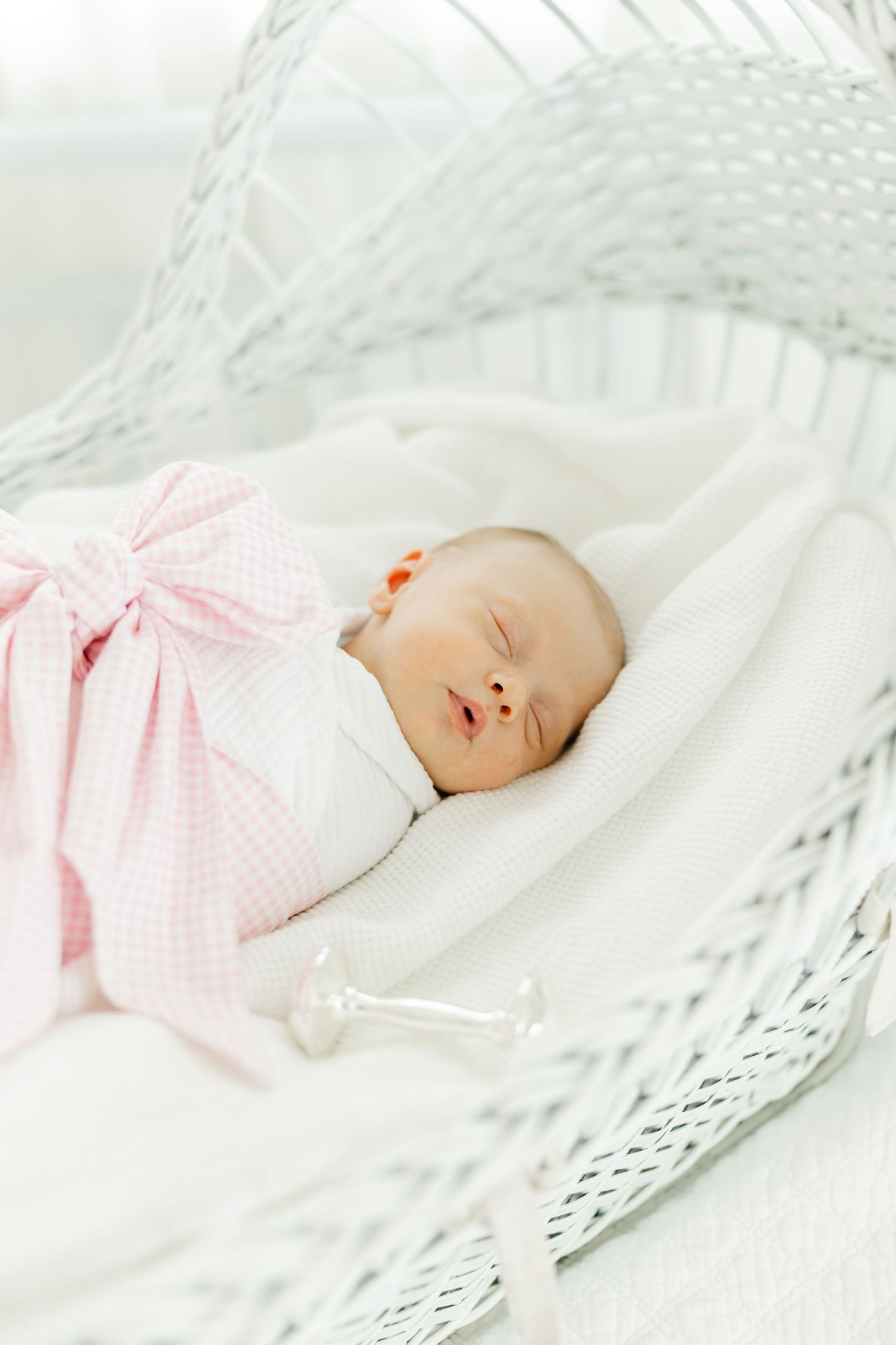 Sleeping newborn baby wrapped in a pink gingham swaddle with a bow, resting in a white wicker bassinet.