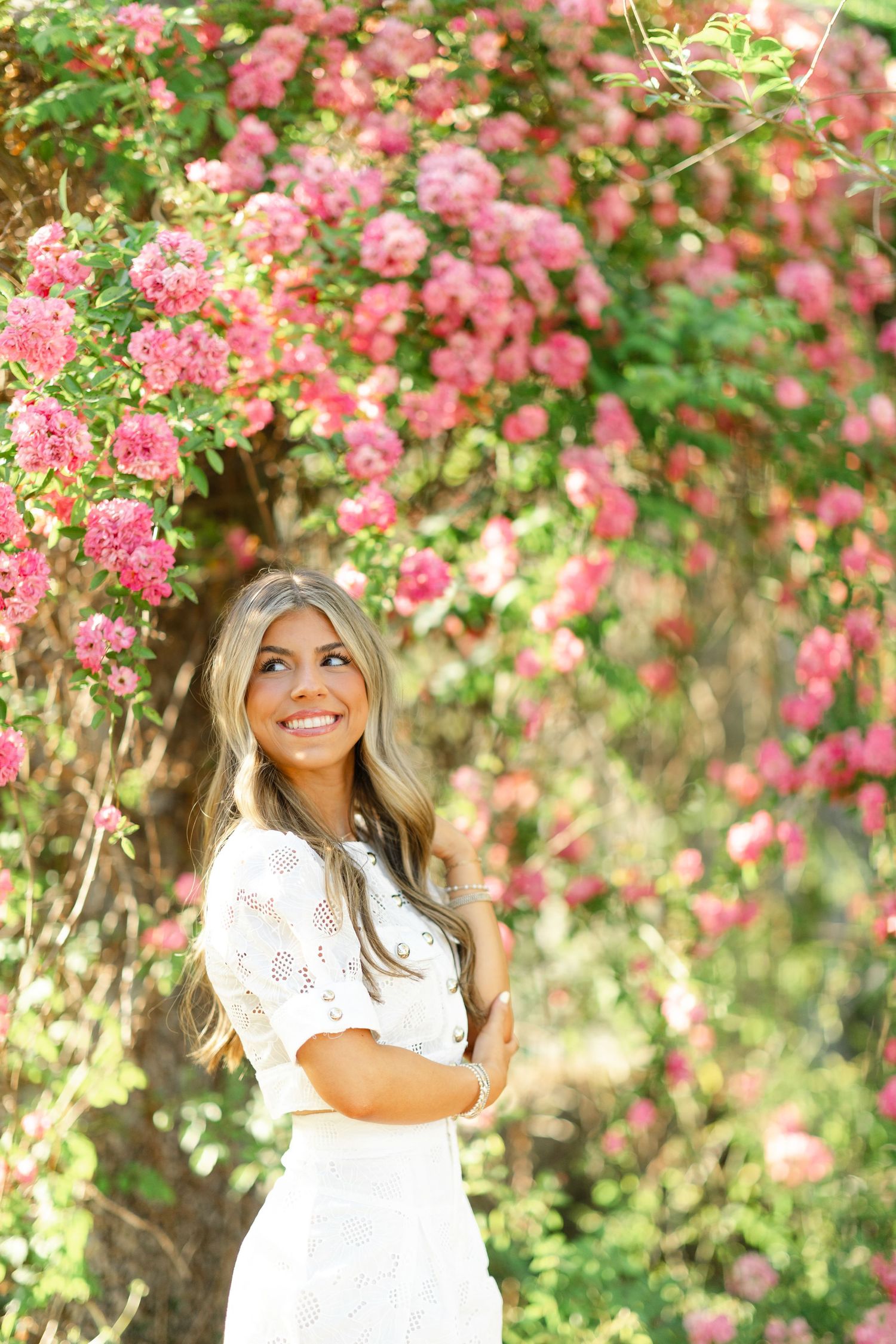 Smiling young woman in white lace dress stands before a vibrant wall of pink climbing roses.