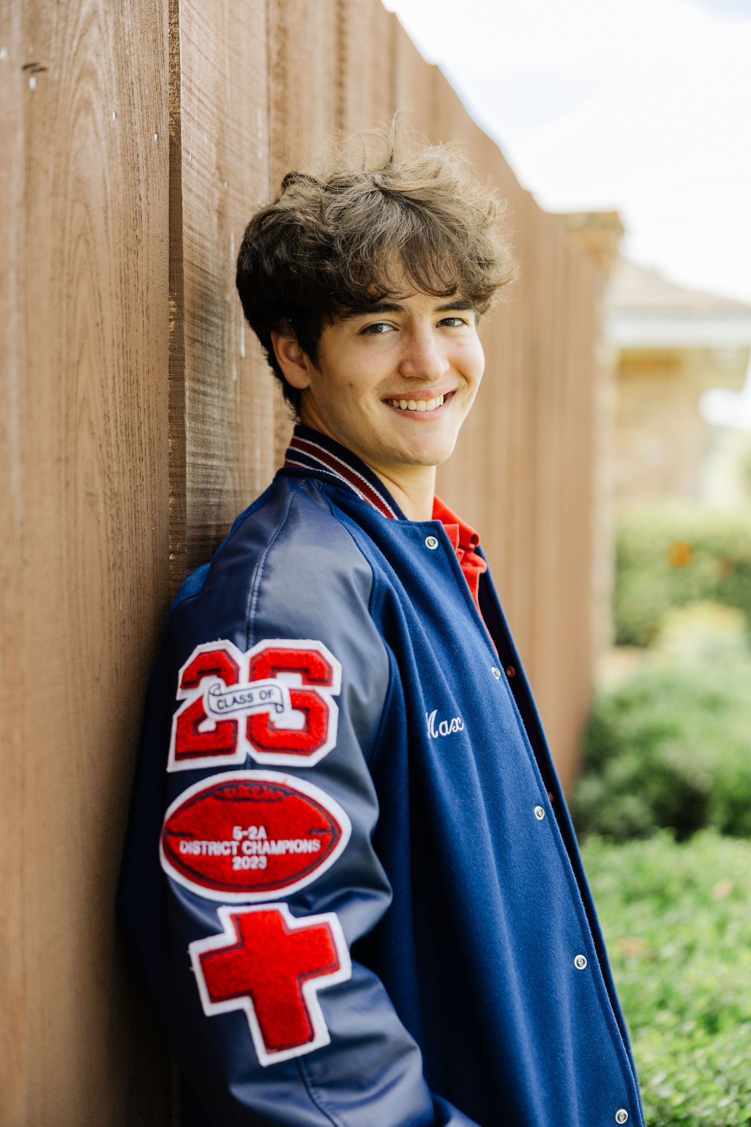 Smiling teenage boy in blue varsity jacket leans against wooden fence for senior portrait photo.