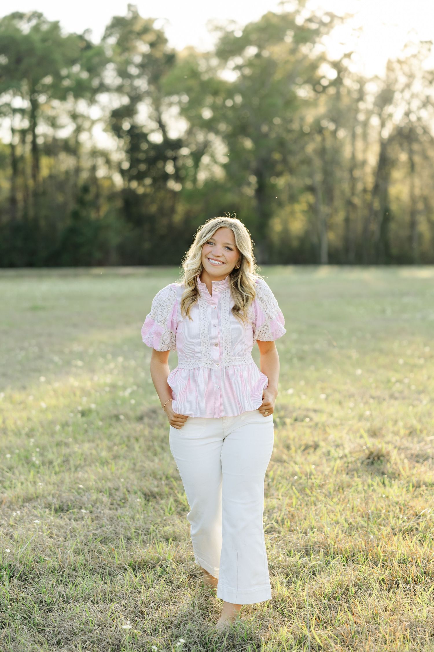 Smiling blonde woman in pink puffed-sleeve top and white jeans poses in a sunlit green field.