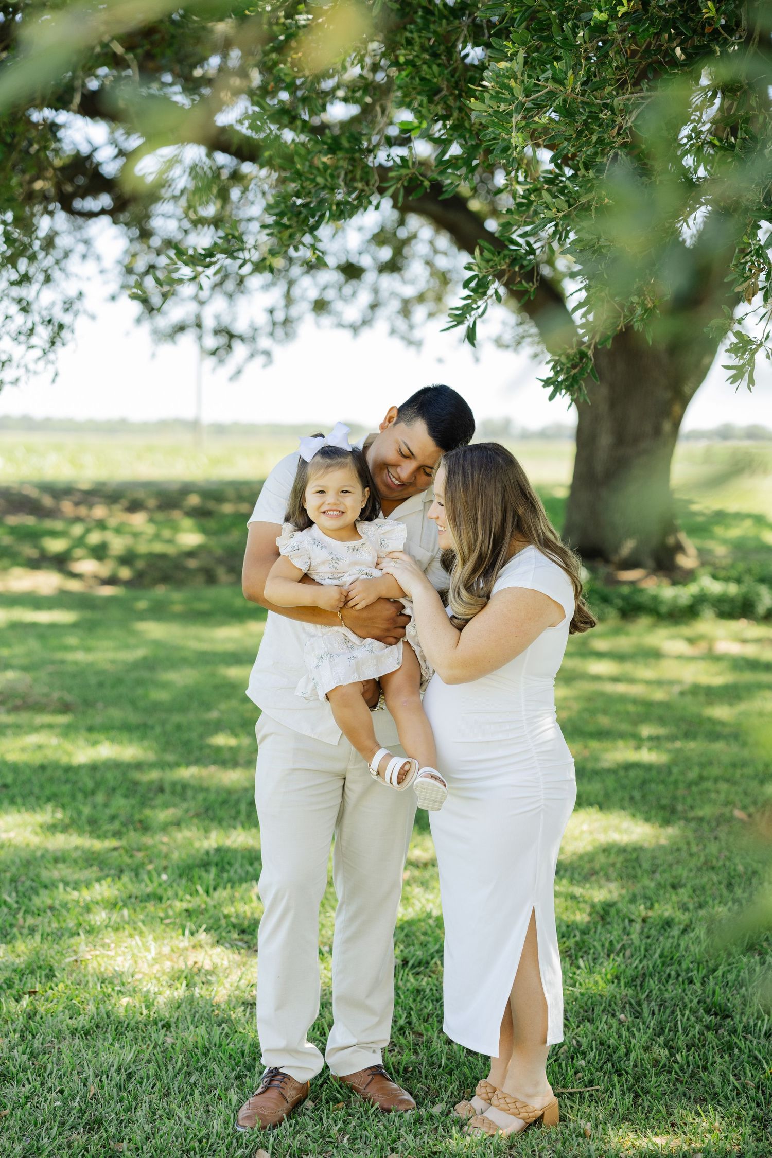 Expecting parents kiss toddler daughter outdoors under large oak tree.
