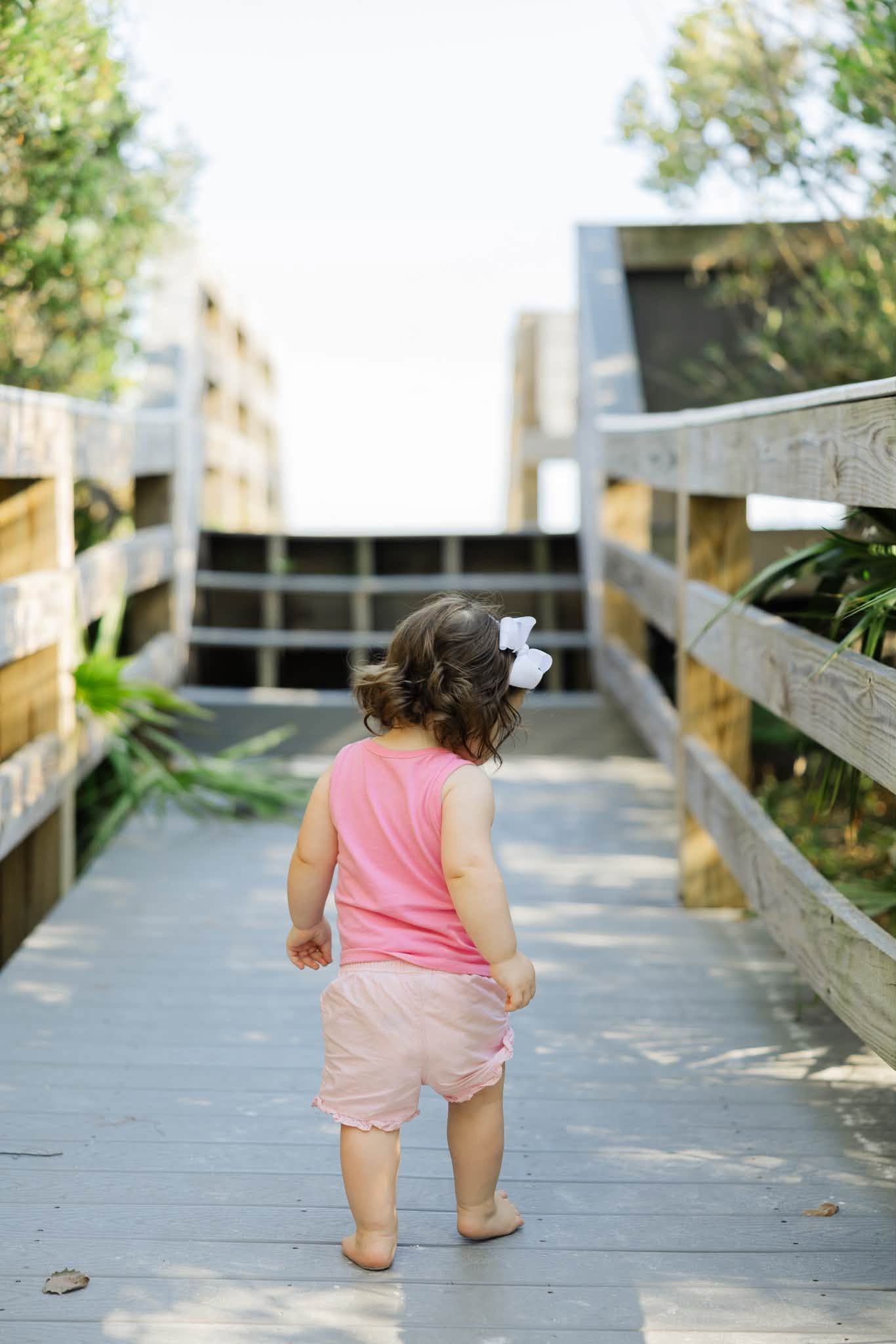 A toddler girl in a pink outfit with a white bow walks along a sunny wooden boardwalk.